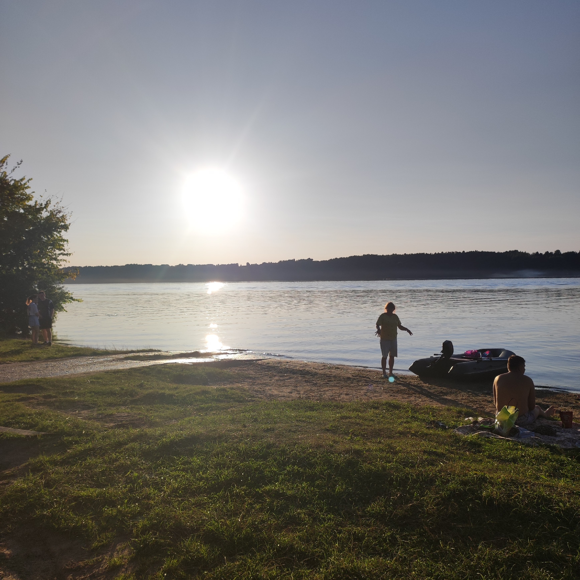 The image captures a serene scene by a body of water, likely a river or lake, on a bright, sunny day, with the sun positioned high in the sky and casting a gentle glare across the water's surface. The foreground is dominated by lush green grass, with a dirt path leading towards the water.
Along the sandy shoreline, several individuals are present. To the far left, two figures stand near a large tree. Further down the beach, a person in a yellow shirt and shorts stands facing the water, appearing to be interacting with a dark-colored inflatable boat equipped with a motor. Another individual is seated on the sand next to the boat, facing away from the camera. A light-colored bag and some containers are scattered around the seated person.
In the mid-ground, the wide expanse of water reflects the sky and the sun, creating a shimmering effect. The opposite bank of the water is lined with a dense forest. The overall atmosphere is tranquil and suggests a leisurely outing by the water. The location is identified as the city of Navoloki, Russia.