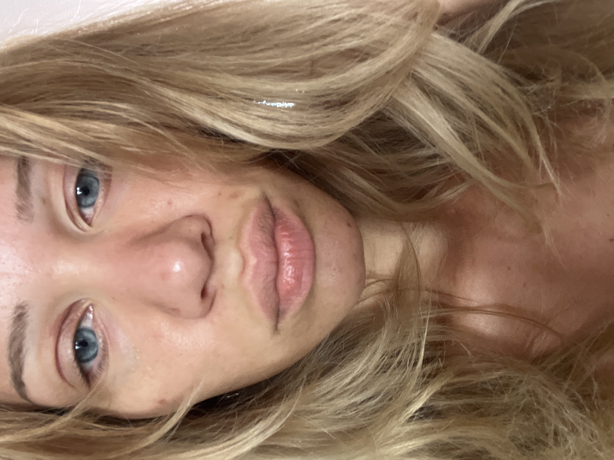 A close-up, eye-level, frontal medium shot shows the face of a young blonde woman with fair skin, light freckles, and blue eyes. Her hair is parted to the left and falls in loose waves around her face. She is looking directly at the camera with a neutral expression. The background is a plain, off-white wall, suggesting an indoor setting. The image does not provide any specific location information that would allow for pinpointing it to Mühldorf a. Inn, Germany.
