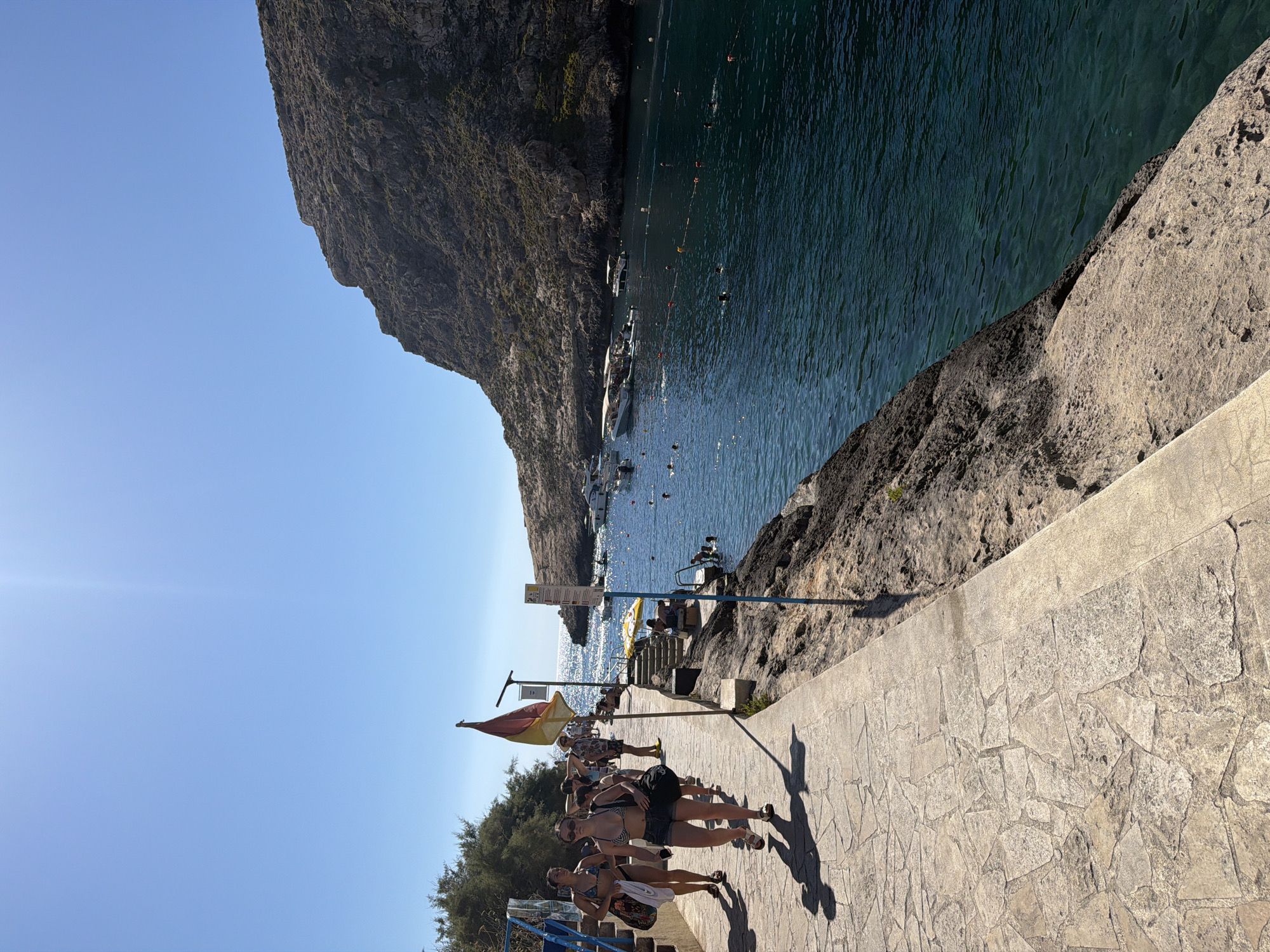 The image depicts a sunny day in Xlendi, Malta, with a picturesque view of the bay. A paved walkway lines the shore, bordered by rocky terrain. Several people are walking on the path, dressed in swimwear, suggesting they are heading to or from the water. The bay is dotted with boats and numerous individuals swimming and enjoying the clear blue water. A red and yellow flag, possibly a warning or information flag, is visible on a pole near the walkway. The background features a steep, rocky cliff face characteristic of the Maltese coastline, under a bright blue sky. The scene conveys a lively and recreational atmosphere at a popular coastal destination.