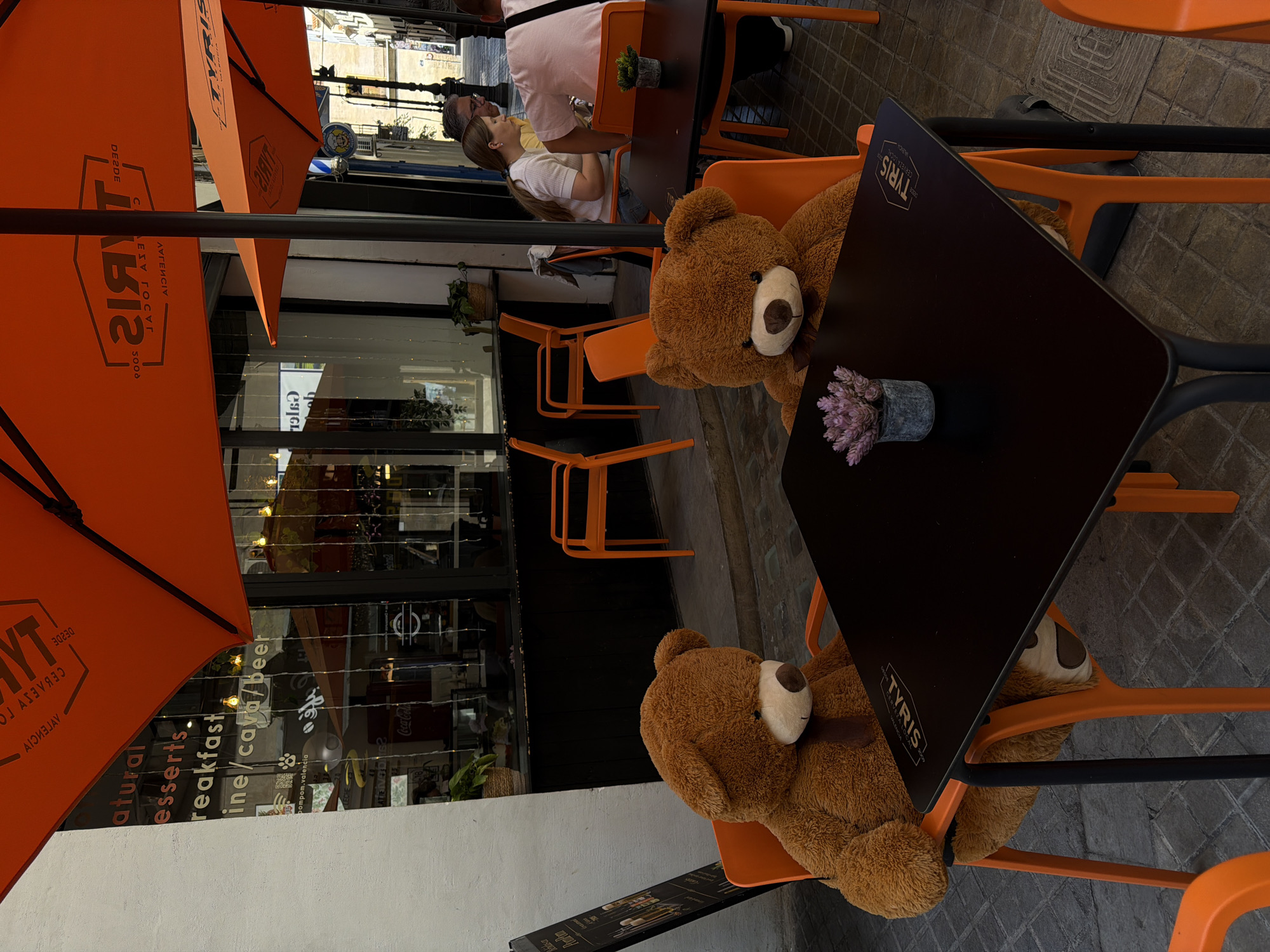 The scene depicts two teddy bears sitting at a table in the outdoor seating area of "TYRIS Cerveza Local" in Valencia, Spain. One bear is seated at the top of the frame and the other at the bottom, facing each other across the table. The chairs are orange. A pot with purple flowers sits on the dark tabletop between them. An orange "TYRIS" branded umbrella shades the area. In the background, a person is sitting at another table, wearing a white shirt and a face mask. The restaurant's glass front displays advertising text like "natural, desserts, breakfast, wine/cava/beer" and the words "pompom.valencia" and "Coca-Cola."