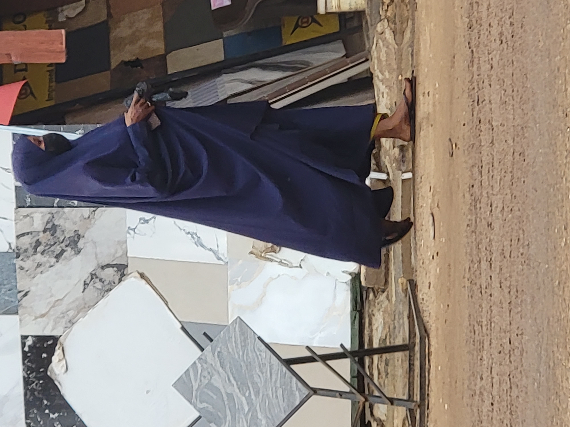 This image, taken in Maiburiji, Nigeria, depicts a person standing in an outdoor setting, likely a market or a construction material display area, on a bright, clear day. The individual is dressed in a dark blue, loose-fitting garment that covers most of their body, including a head covering that obscures their face. Their right arm and hand are visible, holding a dark-colored object resembling a mobile phone or a small camera. Bare feet, wearing simple sandals, are also visible.
The scene is dominated by a variety of tiles or stone slabs displayed around the person. These include square and rectangular pieces with different patterns such as marble-like veins, plain white, and textured grey, some of which appear to be stacked or arranged on a black metal display rack. A small metal-framed table with a square, patterned top, possibly another tile sample, stands in the foreground.
The ground to the right of the person is unpaved, dusty, and sandy, characteristic of an outdoor street or pathway. In the background, various items like wooden planks and cardboard boxes are visible. A yellow cardboard box in the upper background features a black logo and partially visible text that includes the word "improved" and "LO" or "1.0". The person's activity is not explicitly clear, but they appear to be standing, possibly observing the surroundings or interacting with the object in their hand. The bright illumination indicates it is daytime, and the dusty ground suggests dry weather conditions.