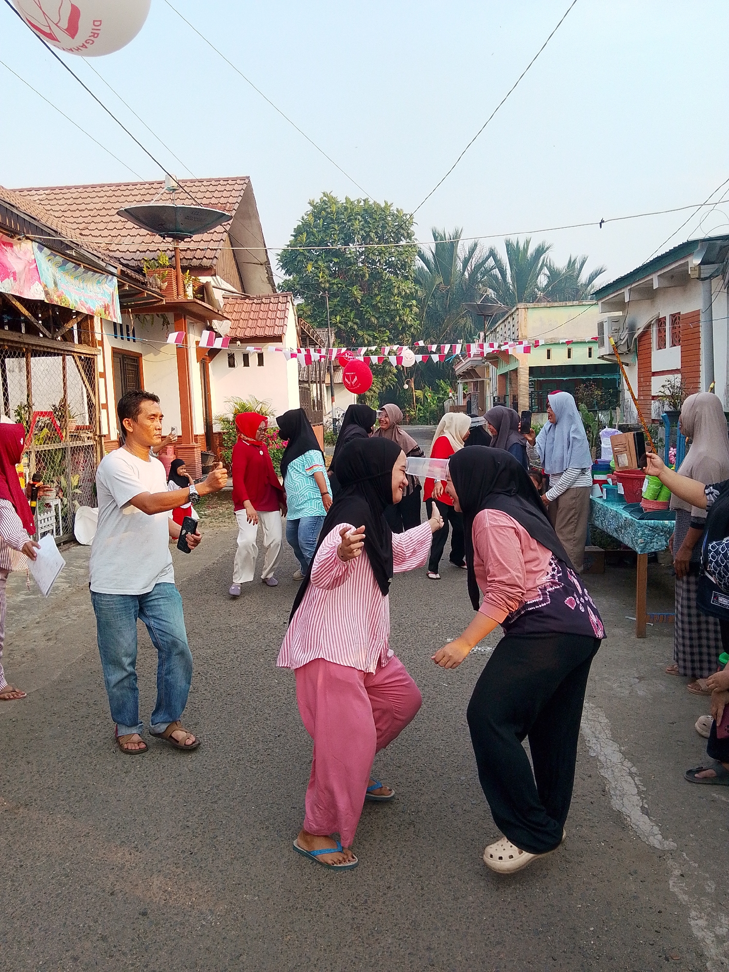 The image depicts an outdoor gathering on a paved street in Barabai, Indonesia, likely for a local celebration. The scene is characterized by a modest residential setting with houses lining the street, adorned with red and white Indonesian flags and pennant banners, suggesting a national holiday or event. A group of people, predominantly women wearing hijabs, are engaged in what appears to be a recreational activity or game. One woman in a pink and white striped shirt and pink pants is dancing or moving dynamically, facing another woman in a black hijab and patterned shirt who is also in a playful stance. Other individuals are observing, interacting, or participating in their own ways; one man in a white t-shirt and jeans is holding a phone and gesturing. A table laden with various containers, possibly for a game or refreshments, is set up on the right side of the street. The overall atmosphere seems festive and communal, with the warm, natural lighting indicating it is daytime, likely late afternoon given the angle of the sun.