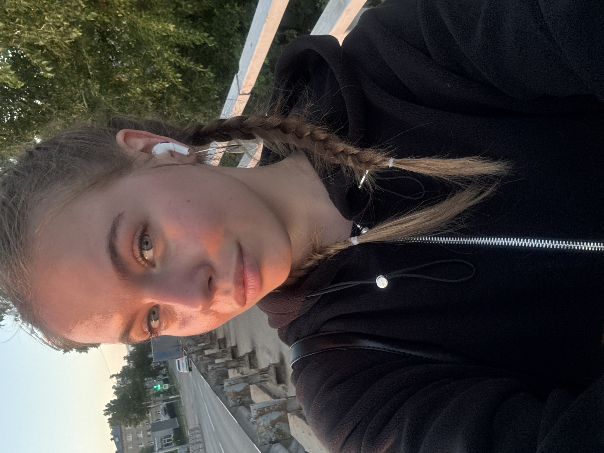 The image is a rotated selfie of a young person, likely female, taken outdoors in what appears to be Chaikovsky, Russia. The subject has fair skin, light brown hair styled in two braids secured with white hair ties and silver-colored clips, and light-colored eyes. She is wearing white in-ear headphones (AirPods) in her left ear and a black hooded fleece jacket with a silver zipper. A black bag strap is visible over her shoulder.
The setting is an outdoor urban or suburban environment. The background shows a road with concrete barriers and a white railing, suggesting the person might be on a bridge or overpass. Further down the road, buildings, a white car, and a green traffic light are visible. Green trees with dense foliage are in the upper left of the frame.
The activity appears to be the subject taking a self-portrait while possibly listening to audio through her headphones. The notable details include the warm, golden lighting on her face and the sky, which indicates late afternoon or early evening, possibly sunset. The weather appears clear. The subject's expression is neutral and calm. There is no visible text in the image.