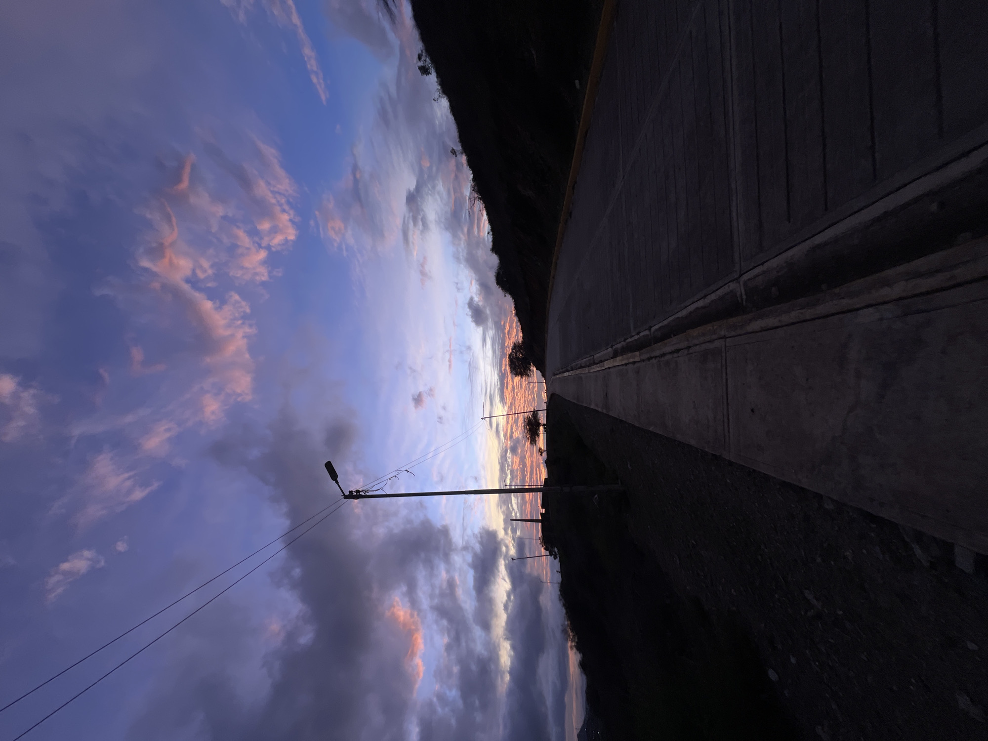The image depicts a road and sidewalk on a hillside during twilight, with a dramatic sky filled with pink and purple clouds. A silhouette of a street lamp and power lines are visible on the left side of the frame. The road curves to the right, ascending the hill, with a dark hillside on the right and a dark valley with some distant lights on the left. The scene appears to be in a residential area on the outskirts of Santa Ana, Venezuela, likely overlooking the city. The overall impression is one of a serene and picturesque evening.