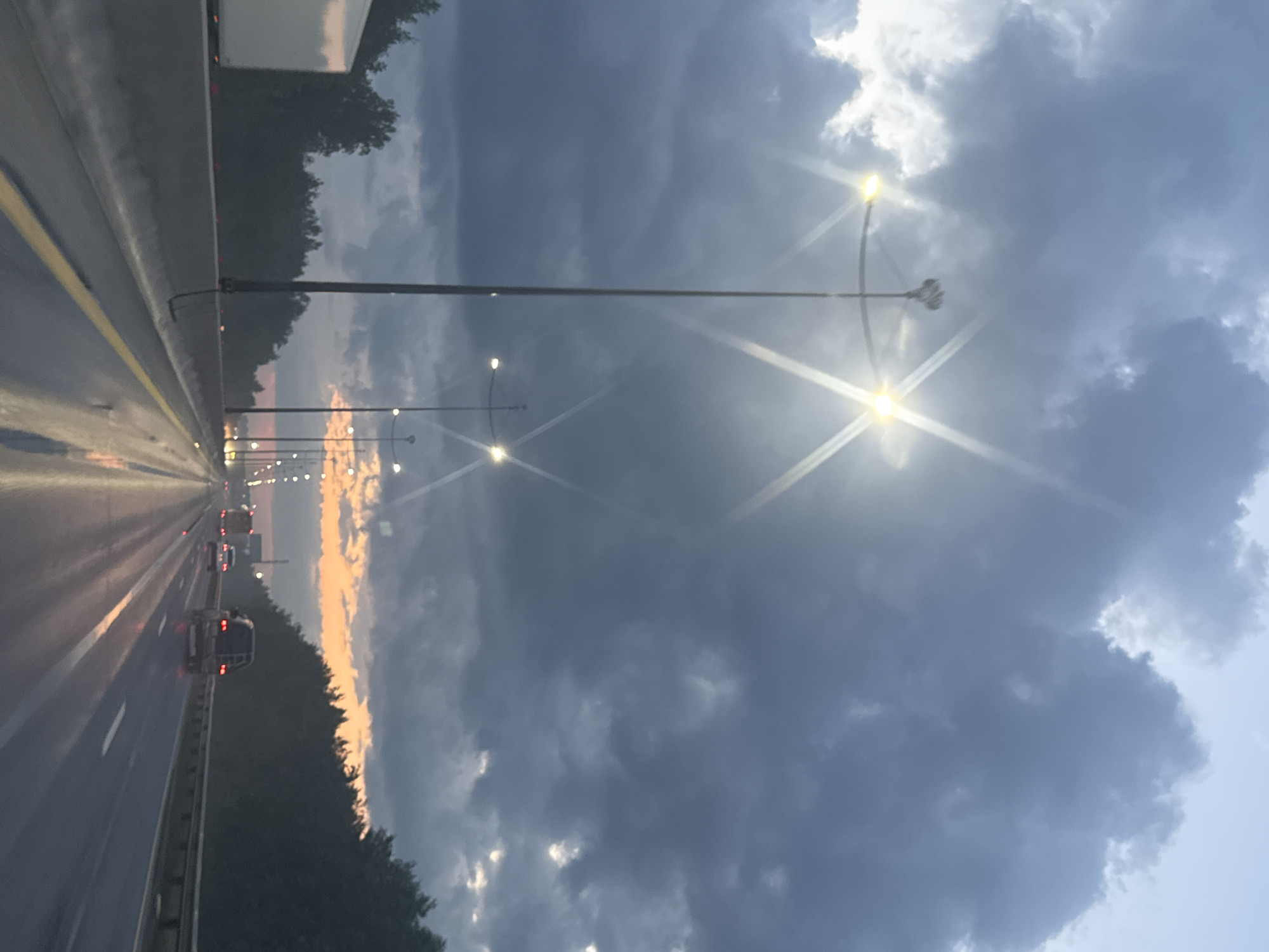 The image depicts a highway in Kondratovo, Russia, during a cloudy dusk. The sky is filled with dark, dramatic clouds with patches of lighter sky peeking through. Streetlights with bright, star-like flares illuminate the highway. Multiple vehicles are visible on the road, including a white truck on the left, a dark SUV in the right lane, and other cars in the distance. The wet surface of the highway reflects the lights and the sky. A line of trees borders the highway on the right side, with a forest visible beyond.