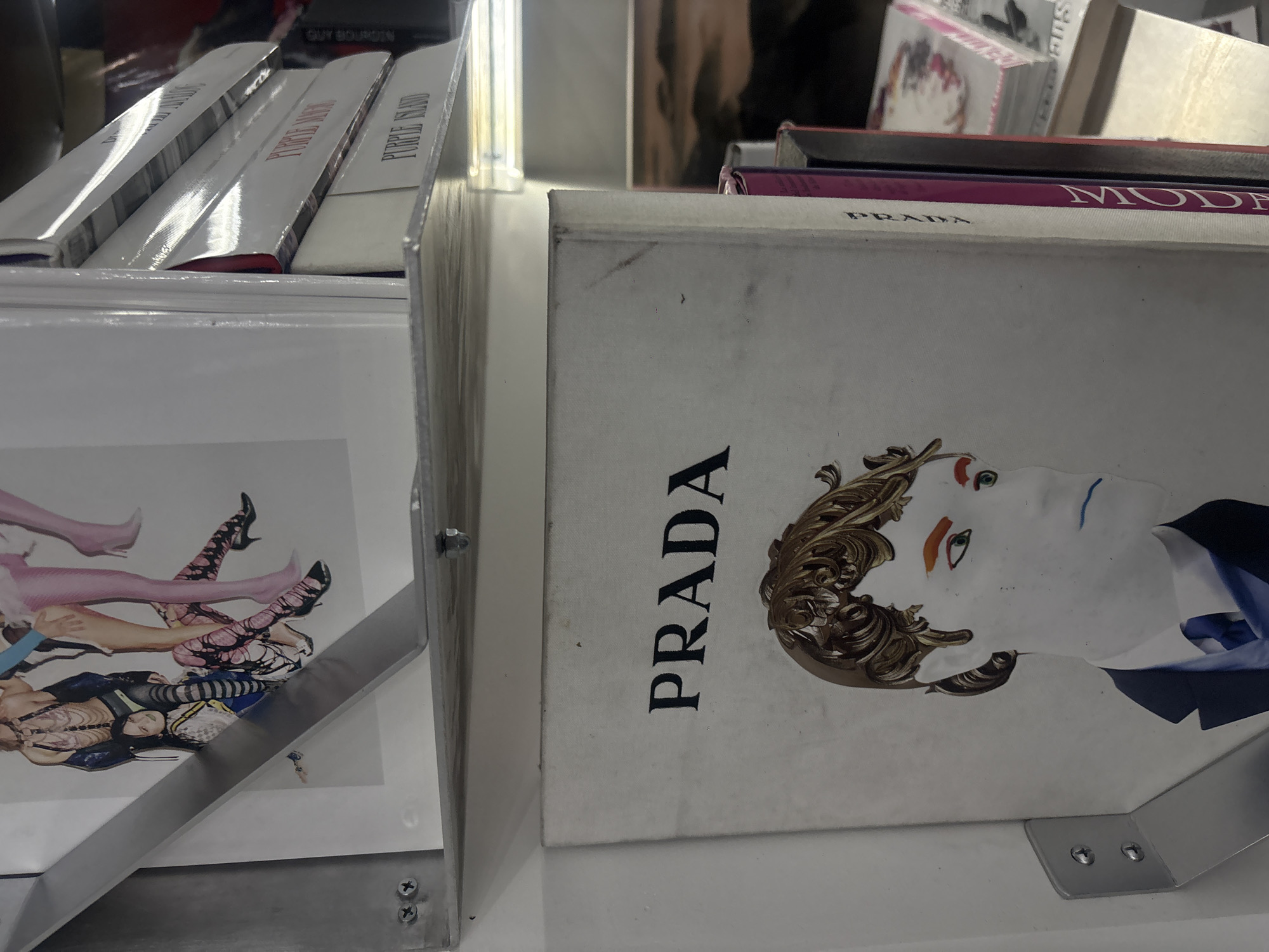 The image shows two books standing upright in metal bookends. One book has the word "PRADA" in bold black letters on the front cover, accompanied by an image of a person's face and ornate hair. The second book has a cover image of multiple legs wearing hosiery and black shoes. The books appear to be situated in a bookstore or retail environment, potentially in Mexico City, Mexico, given the presence of books and bookends. Other books are stacked behind these two, suggesting they are part of a collection or display.