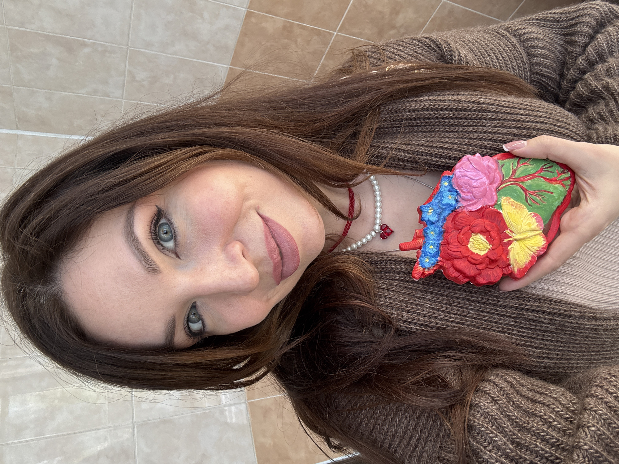 A young woman with brown hair and blue eyes is holding a decorative, colorful object with a floral design in her right hand. She is wearing a brown, knitted cardigan over a beige top and a pearl necklace with a red pendant. The background consists of light brown and beige square tiles. The setting seems to be indoors. There are no explicit indicators to confirm the location as Kryvyi Rih, Ukraine, or any specific city.
