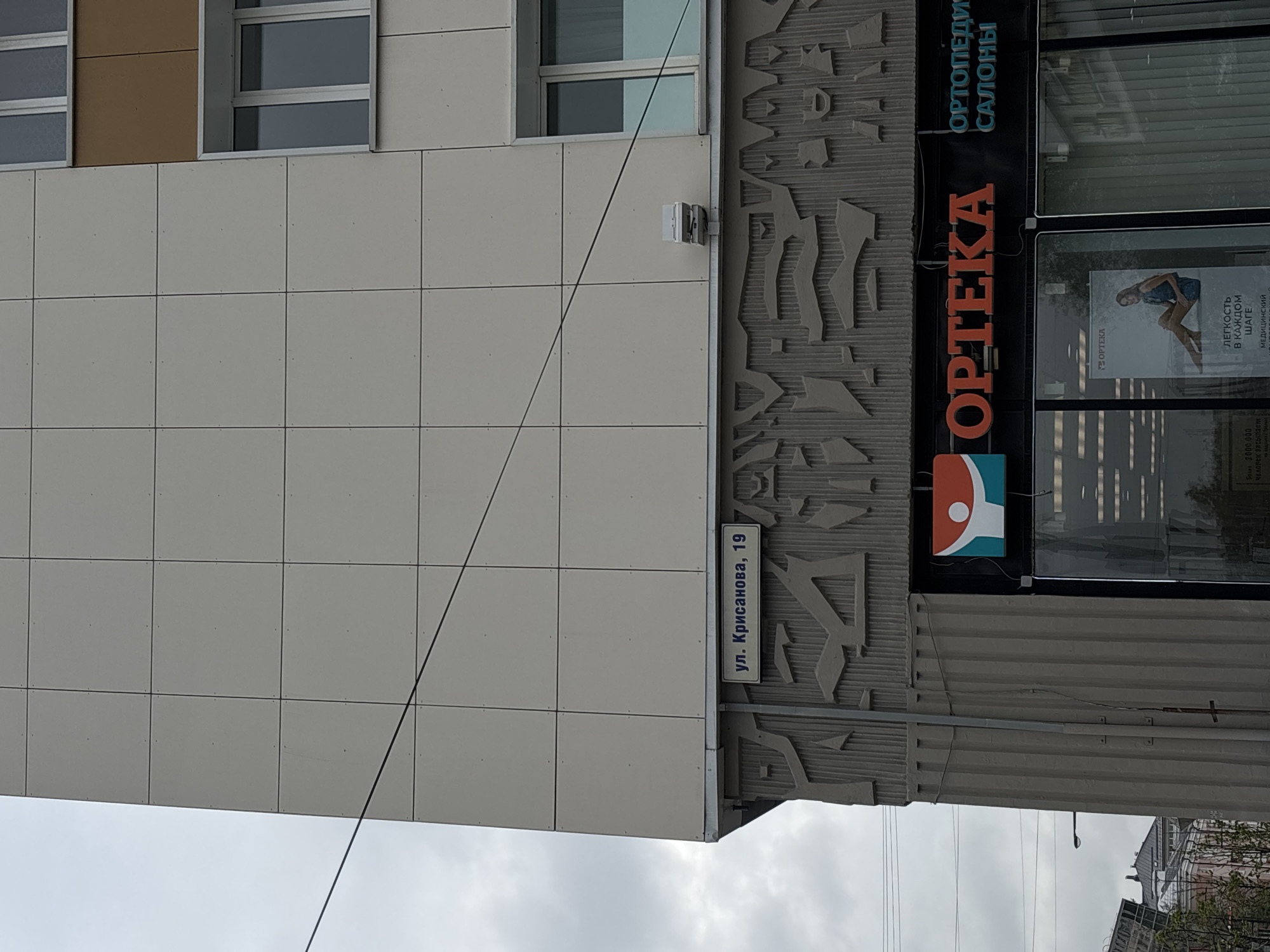 The image shows the exterior of a building in Perm, Russia. The address "ул. Крисанова, 19" (Ulitsa Krisanova, 19) is visible on a sign. There's an "OPTEKA" sign, which likely indicates a pharmacy. Another sign says "ОРТОПЕДИ САЛОНЫ" (Ortopedi Salony), indicating an orthopedic salon. A poster advertising "OPTEKA" is visible in a storefront window. There are powerlines overhead. The building facade is primarily covered with square white or light grey panels, with windows visible higher up. The sky is overcast.