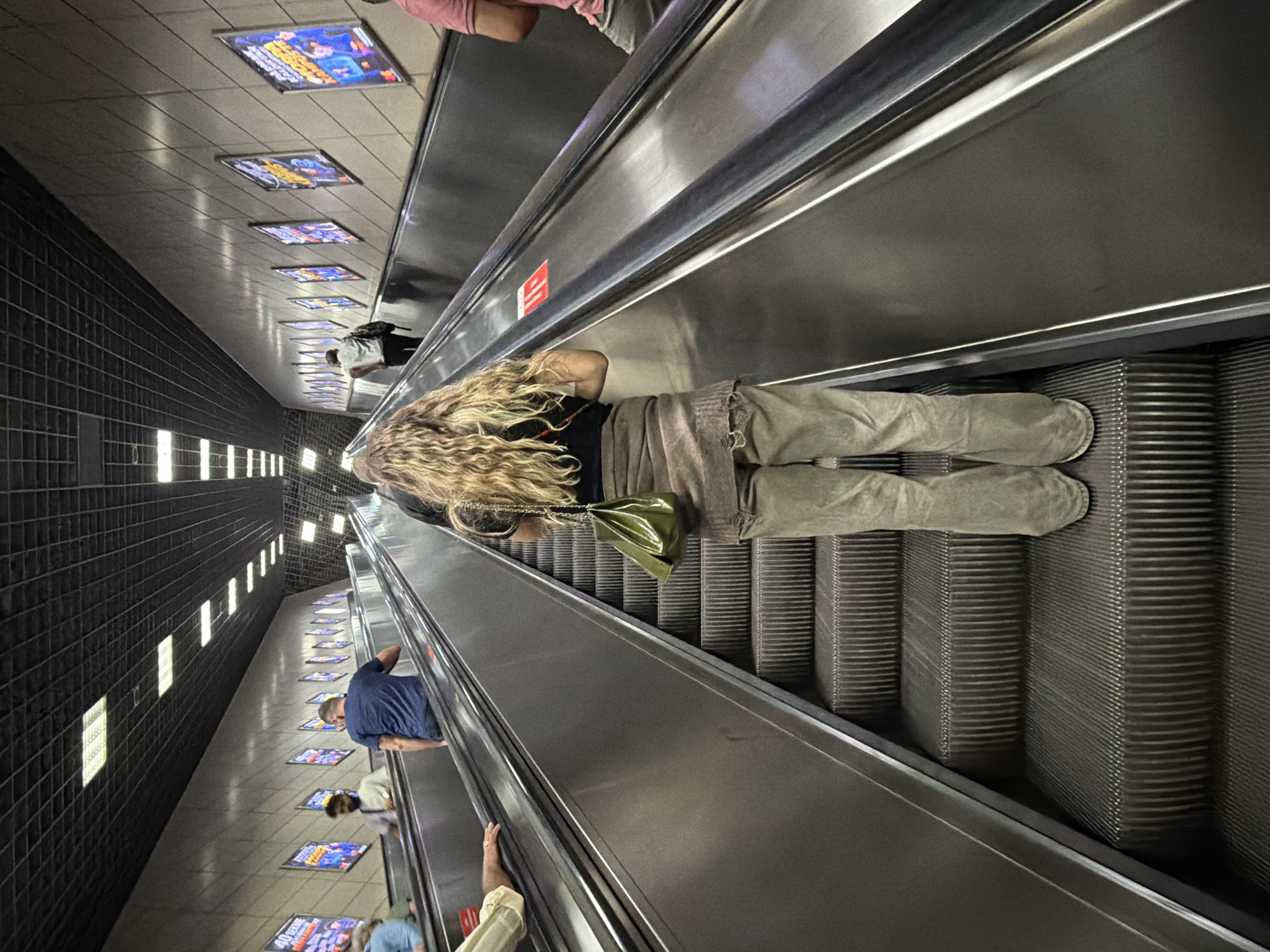 An outdoor escalator ascends a slope in Istanbul, Turkey, likely within a public transportation hub given the overhead lighting and wall-mounted advertisements. The escalator's metal stairs are visible, some worn from use, as are the side rails. Several people are on the escalator, mostly moving upwards. A woman with long, wavy blonde hair is in the foreground, facing away from the camera, wearing a black top, a brown shawl or sweater draped over her shoulders, and loose-fitting olive-colored pants. She carries a small, shiny green bag over her left shoulder. To her left, on another escalator going in the same direction, a person in a light-colored shirt and dark pants is visible from the waist down. To her right and further up, a person in a pink polo shirt and light-colored pants is ascending. In the background, other individuals are on escalators and walking. The wall on the right side of the escalator is tiled and features several framed advertisements. The ceiling is a grid of black panels with integrated lighting.