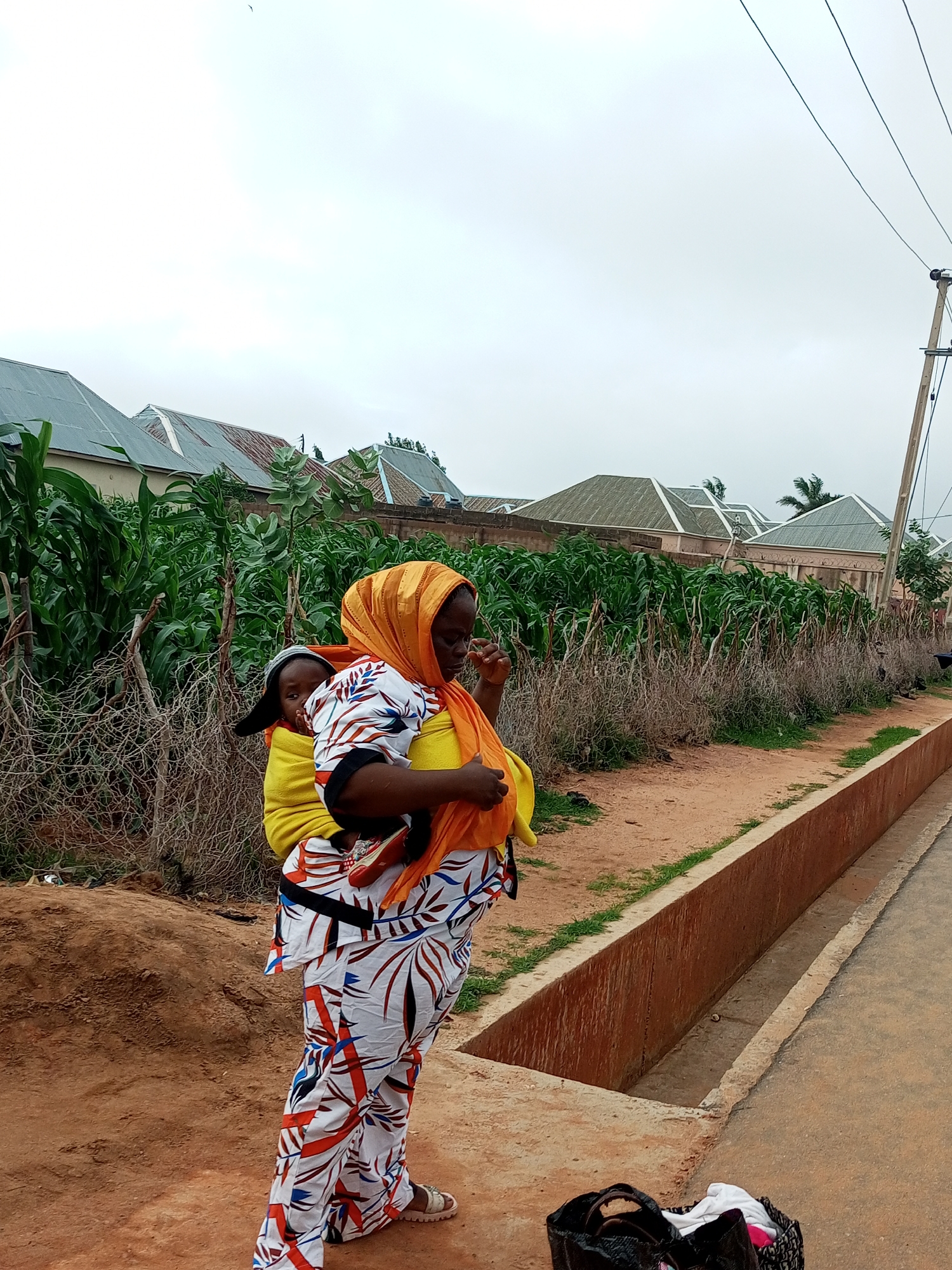 A full-shot, medium-low angle, outdoor photograph shows a woman carrying a baby on her back. The woman is wearing a colorful, patterned outfit with an orange headscarf. The baby is wrapped in a yellow cloth. They are standing on a dirt path next to a drainage ditch. In the background, there are rows of corn stalks and houses with corrugated metal roofs. A utility pole with wires runs up the right side of the image. The sky is overcast.
