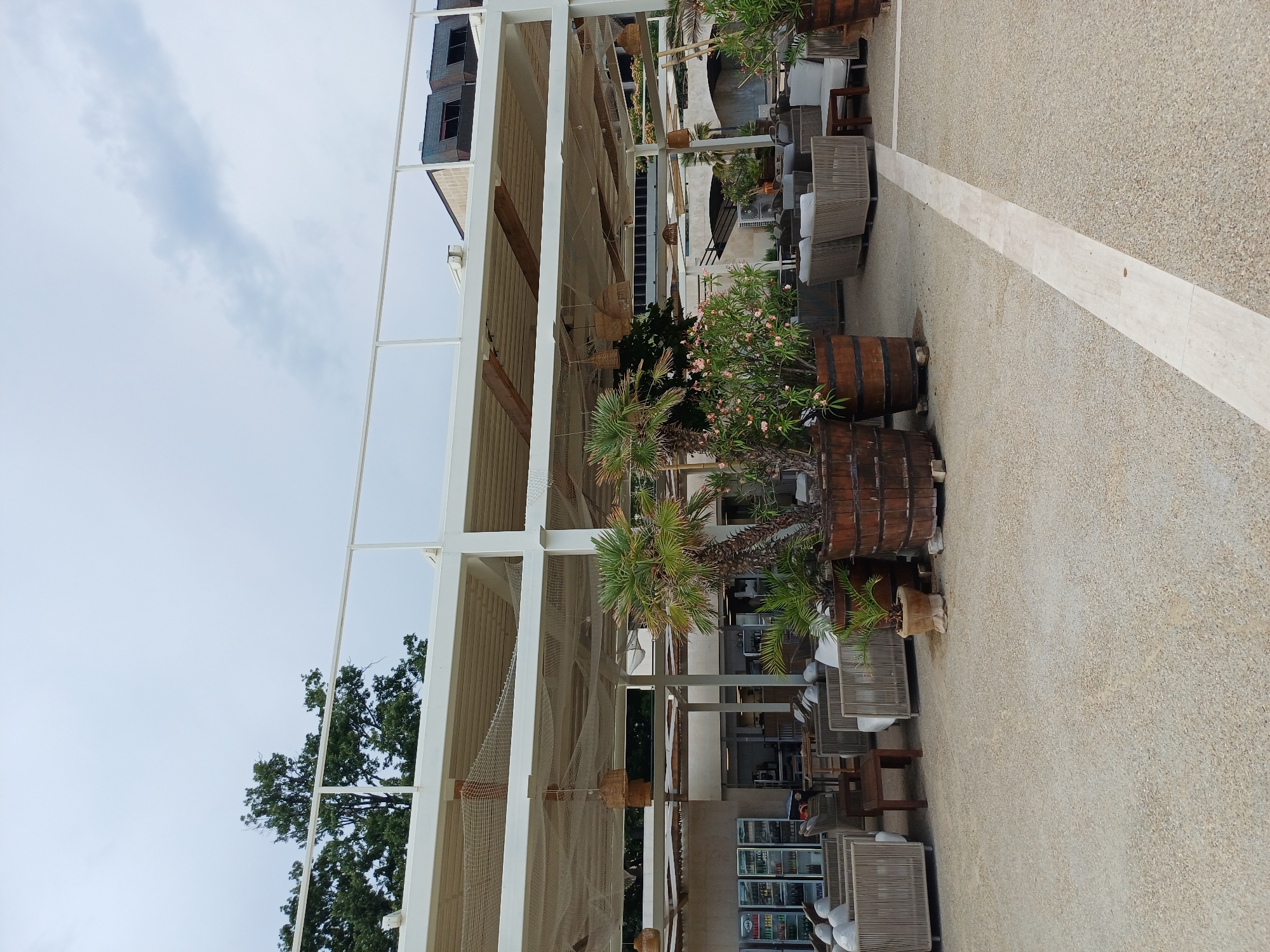 The image depicts an outdoor seating area at a venue located in Varna, Bulgaria. The scene features a covered patio with modern, woven outdoor furniture arranged in conversation groupings. Large wooden planters contain tropical vegetation, including palm trees and flowering shrubs, adding to the ambiance. In the background, a building with a dark roof and windows is visible. Refrigeration units are present, suggesting a bar or restaurant. The ground is covered with a light-colored gravel surface, with a light-colored stone path running through it. The sky is overcast. There are no people visible in the image.