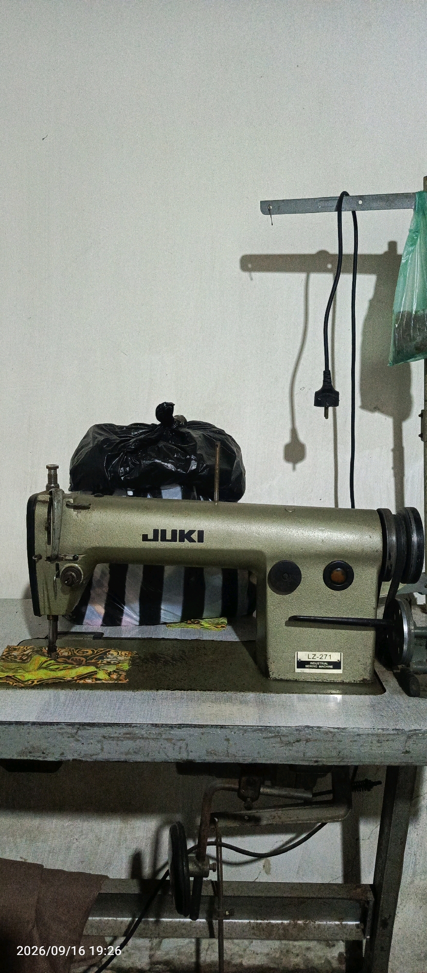 A Juki LZ-271 industrial sewing machine, colored in a muted green, sits on a gray metal table. The machine is positioned to the right of center, with its needle down on a piece of patterned fabric. Behind the machine, a black trash bag is placed on top of the sewing machine's arm. To the right of the trash bag, a black electrical cord hangs from a metal bracket mounted on a white wall. A green plastic bag filled with an unidentifiable dark substance is also hanging from the bracket. The wall itself has some shadows cast upon it, including the shadow of the electrical plug. Below the sewing machine, on the table, is a piece of fabric with a black and white striped pattern visible through the machine's working area. The table appears to be part of a larger workbench structure, with metal supports and a lower shelf containing more mechanical components. The overall scene suggests a workshop or sewing area. The timestamp "2026/09/16 19:26" is visible in the bottom left corner of the image.