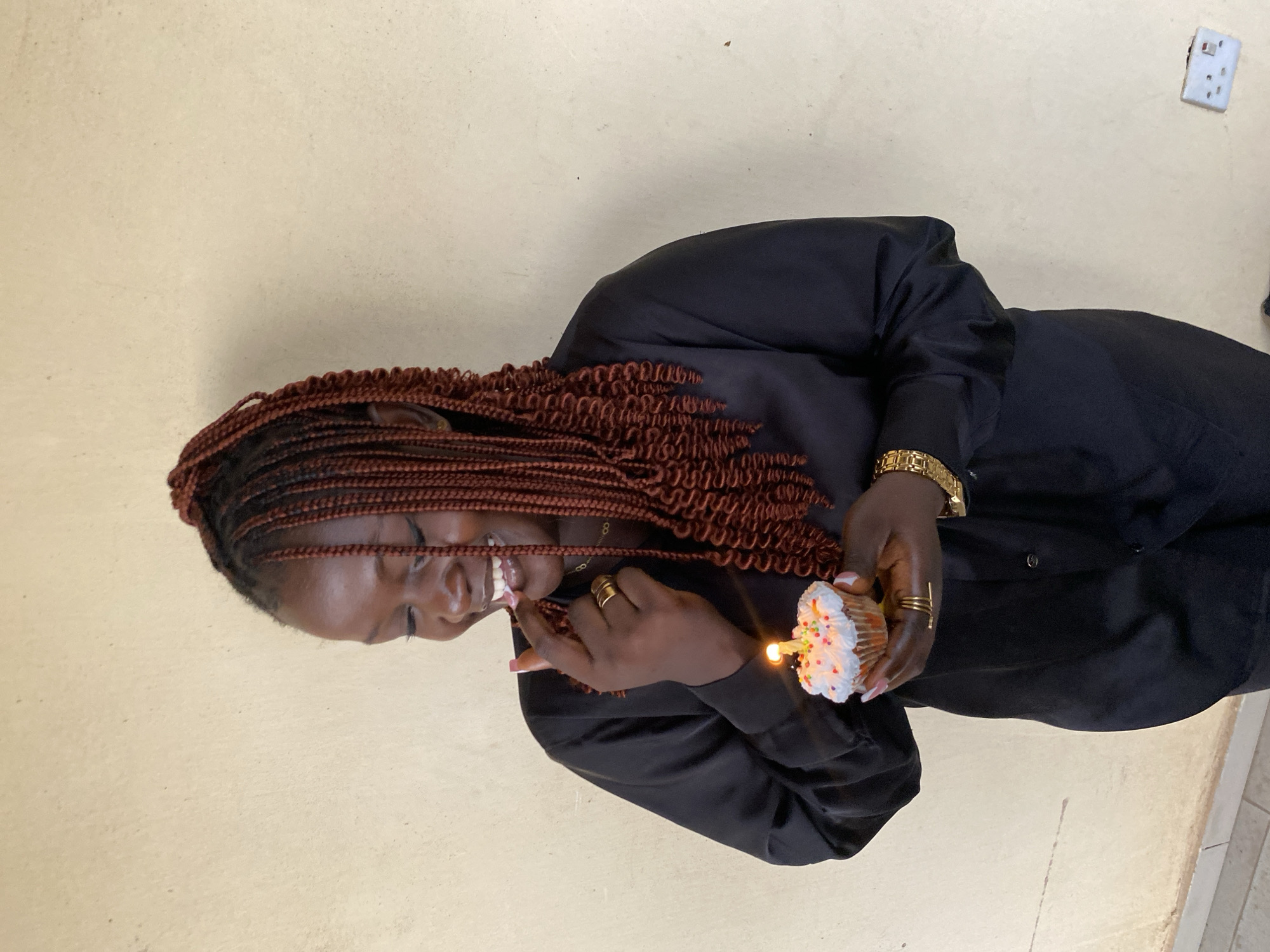 A woman with brown braided hair and wearing a black long-sleeved shirt holds a cupcake with white frosting, rainbow sprinkles, and a lit candle. She is smiling and appears to be licking frosting off her finger. She is wearing a gold watch and gold rings. The background is a plain, light beige wall with a white electrical outlet visible in the upper right corner. While the location is identified as Bukuru, Nigeria, there are no contextual clues in the image to confirm this.