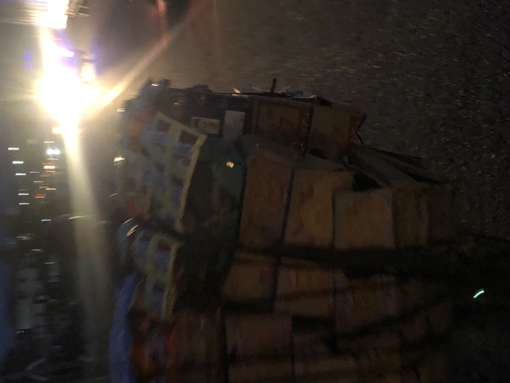 The image depicts a dark, blurry outdoor scene at night in Karu, Nigeria. The primary subject is a large, irregular stack of numerous cardboard boxes, piled high on what appears to be a roadside or ground surface. Many of the boxes show faint branding with yellow, white, and red elements. On some cartons, red text on a yellow background is vaguely visible, potentially resembling a brand name like "Indomie," suggesting food or snack products.
A very bright, overexposed light source dominates the left side of the image, casting a strong glare and creating motion blur. This light could be from a street lamp, vehicle headlights, or an emergency light. Additional faint streaks of light are visible on the far left, possibly indicating other distant light sources or passing traffic.
The arrangement of the boxes, piled seemingly in disarray on a dark ground, hints at an event such as goods having fallen from a vehicle, being offloaded, or involved in some form of roadside incident. No clear human figures are discernible. The overall image quality is poor due to extremely low light conditions, blur, and overexposure from the bright light source.