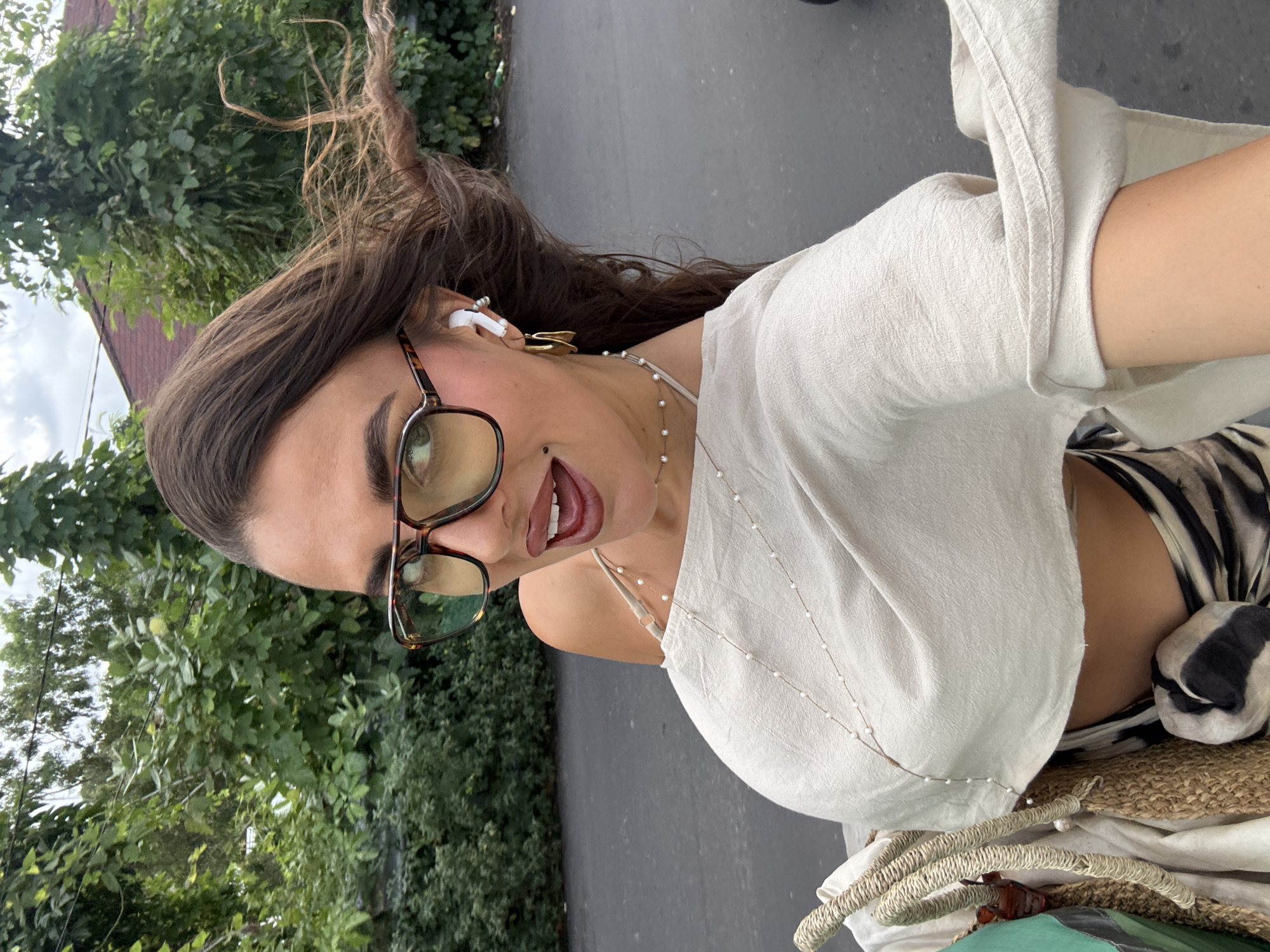A woman poses outdoors. She wears glasses, a white top, and jewelry, including earrings and necklaces. She also has earphones in her ears. The background consists of greenery, foliage, and a grey surface, suggesting a park-like area next to a road. Based on the location provided, Kuta Selatan, Indonesia, the environment suggests an urban area with tropical vegetation. There is no specific information in the image to confirm the exact location or any ongoing actions beyond the woman posing for a photo.