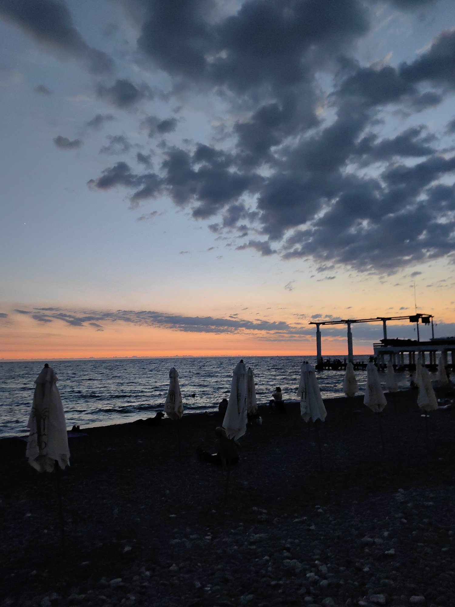 The image captures a serene beach scene at dusk in Sochi, Russia. The sky displays a beautiful gradient of colors, transitioning from a soft pale blue at the top to warm hues of orange and pink near the horizon, indicating sunset. Dark, dramatic clouds are scattered across the sky, adding depth and texture.
The foreground features a dark, pebbly beach with several closed beach umbrellas standing upright. Their white fabric is partially obscured by the low light, giving them a ghostly appearance. Small, silhouetted figures of people can be seen scattered on the beach and in the shallow water, suggesting a leisurely end to the day.
The sea is calm with gentle waves, reflecting the colors of the sky. In the background, a pier or an elevated structure extends into the water, its silhouette stark against the colorful sky. The overall mood is peaceful and tranquil, evoking a sense of calm as day turns into night. The time of day is clearly sunset, and the weather appears to be clear with some cloud cover.