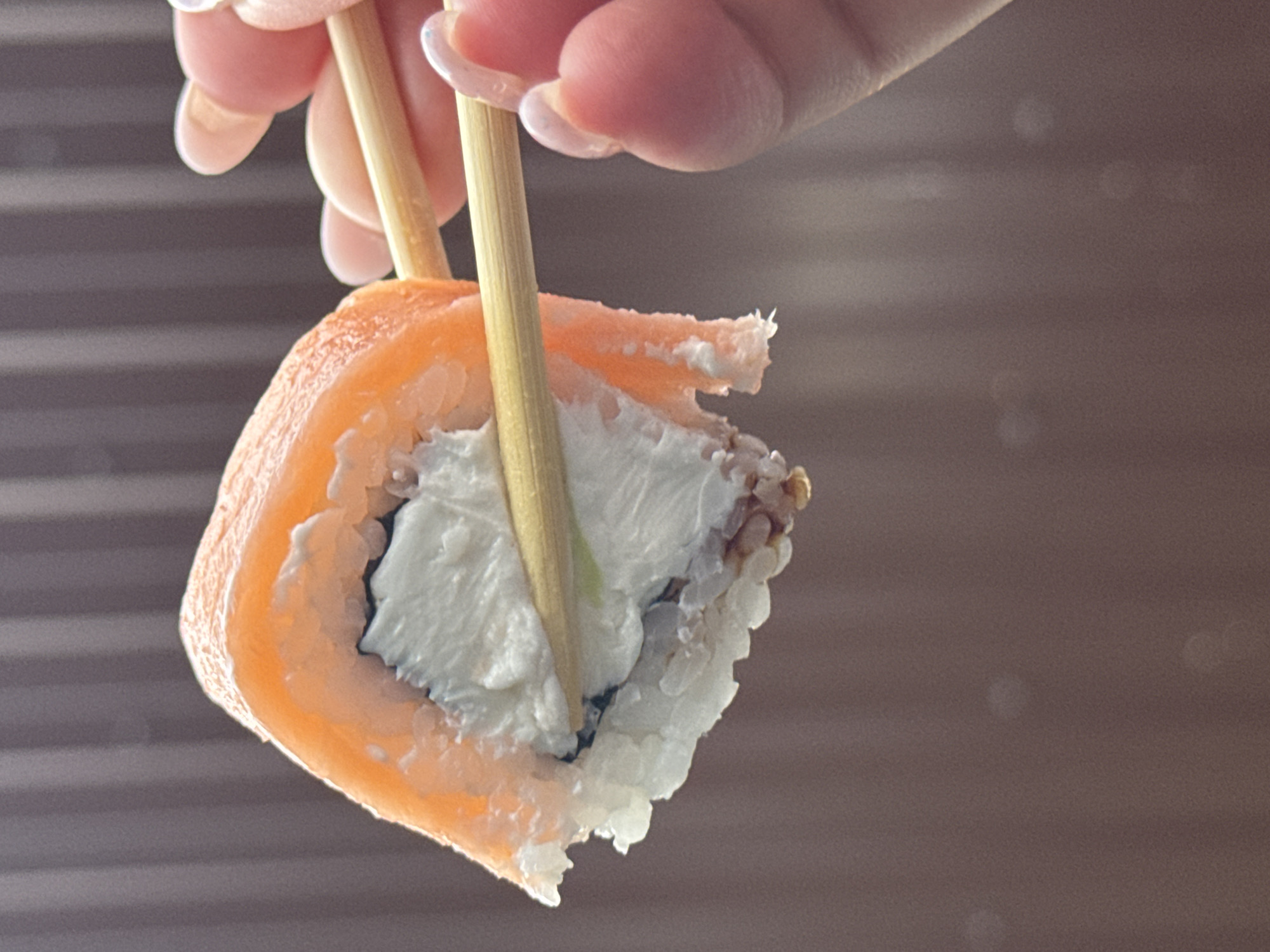 A close-up, vertically oriented, eye-level shot shows a hand holding a piece of sushi with chopsticks. The sushi roll features a bright orange salmon exterior, a white creamy filling, and rice. The chopsticks are made of light brown wood and are inserted into the sushi roll. The hand has light skin with light pink, well-manicured nails. The background is a blurred, neutral tone with vertical lines. There are no identifiable landmarks or elements that can place the image in Mikhaylovsk, Russia, or any specific city. The scene appears to be indoors, possibly in a kitchen or dining area, given the lighting and the focus on the food.