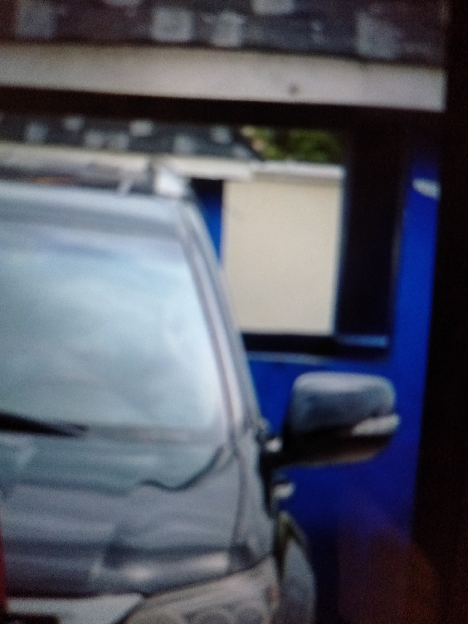 The image depicts a close-up, blurry view of the front passenger side of a dark-colored vehicle, likely an SUV or minivan. The vehicle is parked. The windshield is visible, reflecting a bright, out-of-focus background that includes greenery and structures. To the right of the vehicle, a vibrant blue vertical element is prominent, suggesting it might be part of a building, structure, or possibly a painted surface. A dark gray side mirror is attached to the car door, also out of focus. The lighting appears to be natural daylight. There are no people visible in the frame, and the overall scene lacks distinct features that would pinpoint a specific location within Jalingo, Nigeria.