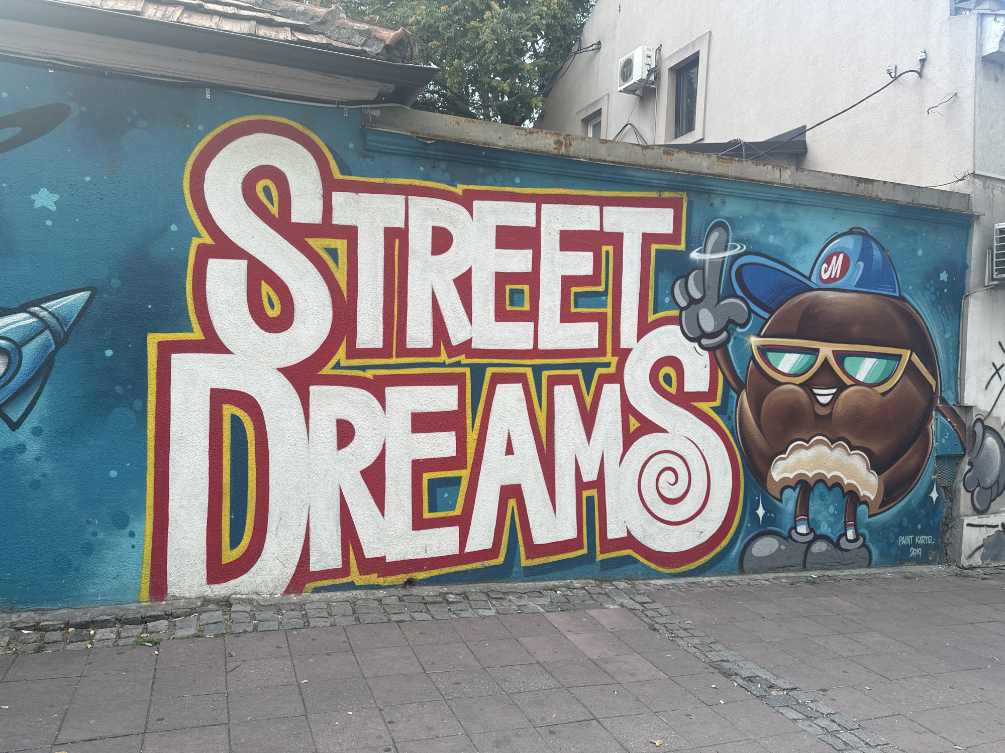 This media file captures a vibrant mural on an exterior wall in Belgrade, Serbia. The mural features large, bold white lettering with red and yellow outlines that spell out "STREET DREAMS." To the right of the text, a cartoon character resembling a coffee bean is depicted wearing a blue baseball cap with an "M" logo, sunglasses, and a fluffy white scarf. The character is raising its index finger, with a circular motion line indicating a gesture.
The background of the mural is a striking blue, with scattered white dots representing stars. To the left of the text, a blue and silver rocket ship is partially visible, adding to a space-themed element. The mural is painted on a textured wall, likely concrete, which appears to be part of a building.
Below the mural, a paved sidewalk with cobblestone edging is visible. The scene appears to be outdoors during daylight, with overcast or diffused lighting, suggesting a mild weather day. The overall impression is one of urban art and creative expression.
Visible text includes "STREET DREAMS" in the mural, and a signature at the bottom right corner reads "PAINT KARTEL 2019". The artistic style is cartoonish and energetic, suggesting a playful and aspirational theme.