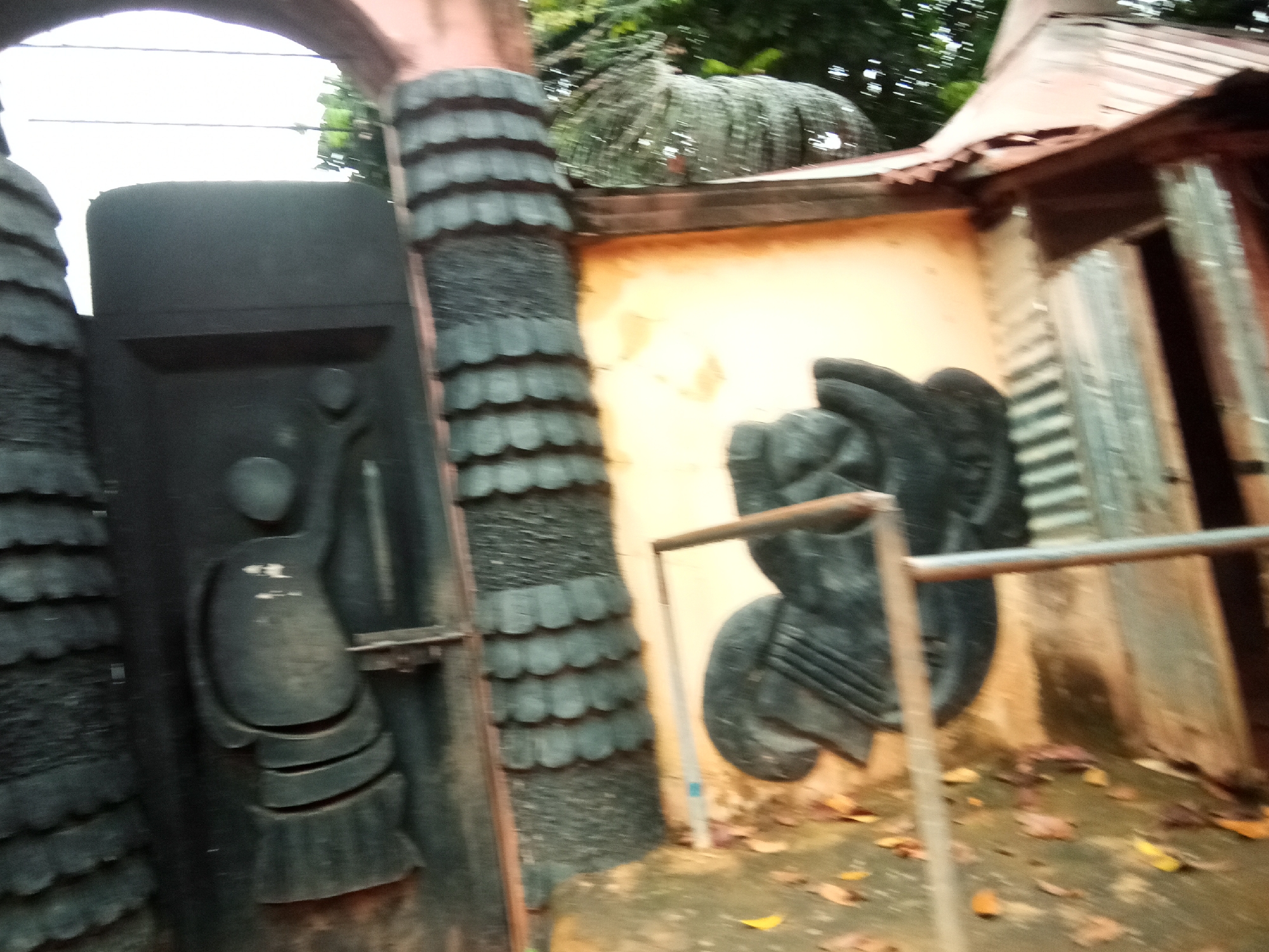 The image shows an outdoor scene in Oko Town, Nigeria. In the foreground, there are two large, dark, sculptural elements. The left element is a vertical structure with layered, scale-like protrusions, resembling a textured pillar or a part of a gate. To its right, a portion of a dark, abstract sculpture is visible, with rounded, layered forms. In the background, a light-colored wall with some texture is present, and a corrugated metal roofline can be seen above it. To the right of the wall, there is an opening with a corrugated metal door. The ground is covered with scattered dry leaves. Trees and foliage are visible in the upper portion of the image. The overall impression is one of an artistic installation or an entryway with decorative, sculptural features.