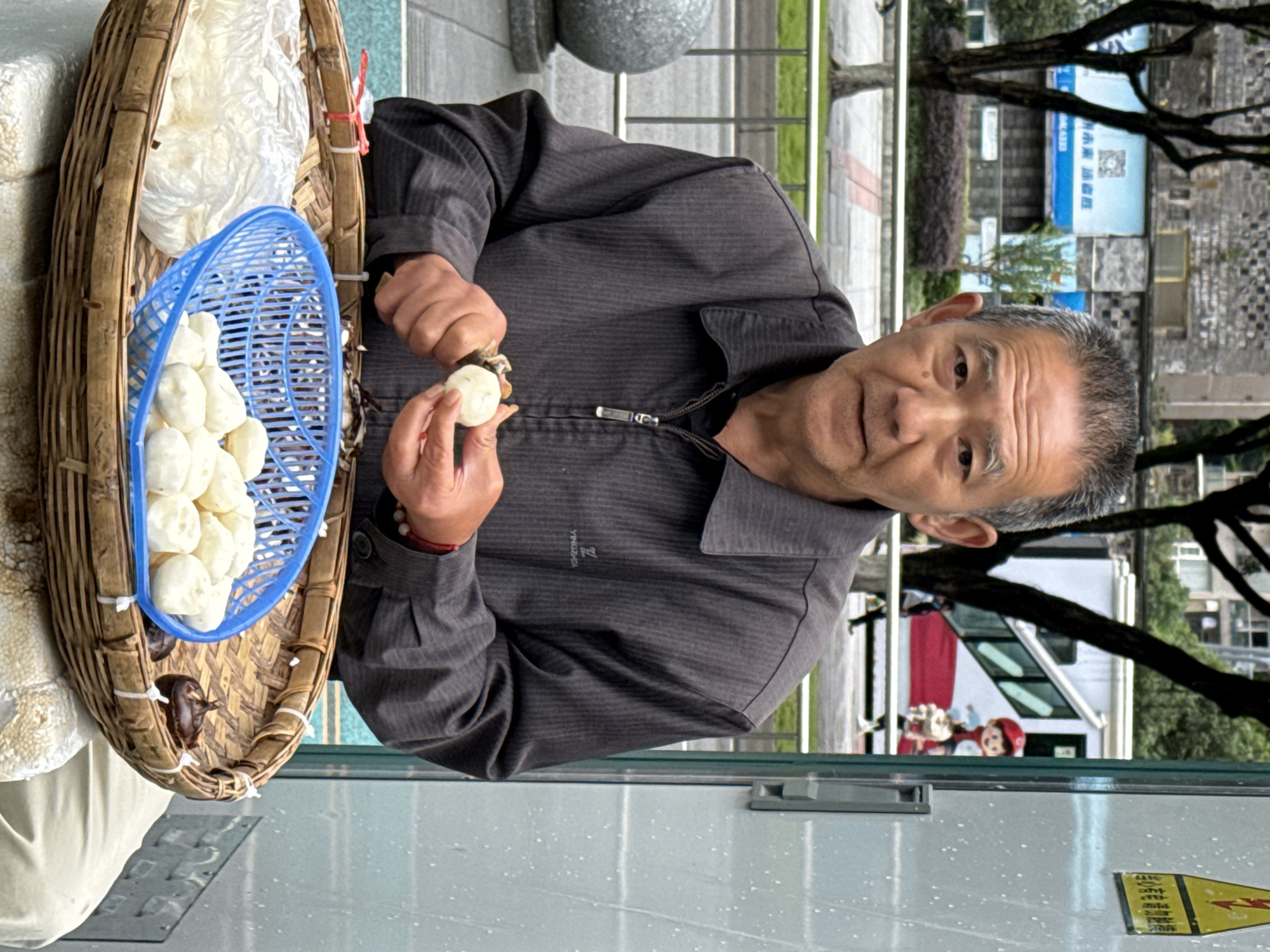 An elderly man is captured in a medium close-up, appearing to be a street vendor, holding and preparing food items. He has short, greying hair and a weathered face, looking directly at the camera with a neutral expression. He wears a dark grey, collared, long-sleeved shirt with a zipper and a red bracelet on his left wrist.
In the immediate foreground, to the man's left, is a large, rectangular bamboo basket, characteristic of those used by street vendors. Inside the basket, a blue plastic strainer holds numerous white, round, steamed food items, likely buns or dumplings. Another plastic bag containing white material is also visible in the basket, along with a few dark, irregularly shaped objects, possibly water chestnuts. The man is holding one of the white food items in his left hand, while his right hand, holding a small tool, appears to be peeling or preparing another one.
The scene is outdoors, likely on a street or sidewalk. The background shows concrete paving, trees with dark trunks, and urban buildings. A blue sign with some Chinese characters and a QR code graphic is visible on one of the buildings. A white vehicle, possibly a van or bus, is partially seen behind the man. The lighting suggests it is daytime. The man and his wares are positioned near what appears to be a light-colored, possibly glass or metal, surface, with a speckled pattern and a partial yellow and red sticker visible in the lower right. On the man's shirt, the brand name "YINGZHEBA" or similar is faintly visible. The image is taken in Jiulongpo District, China.
