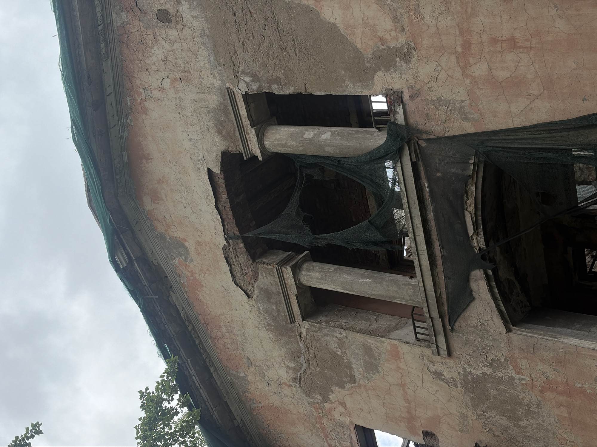 The image shows a portion of a decaying building facade in Saint Petersburg, Russia, featuring crumbling plaster, exposed brick, and structural elements like columns. A dark green safety net is draped over part of the structure, suggesting ongoing construction or preservation efforts. The architecture appears to be classical, although weathered and damaged. The sky is overcast.