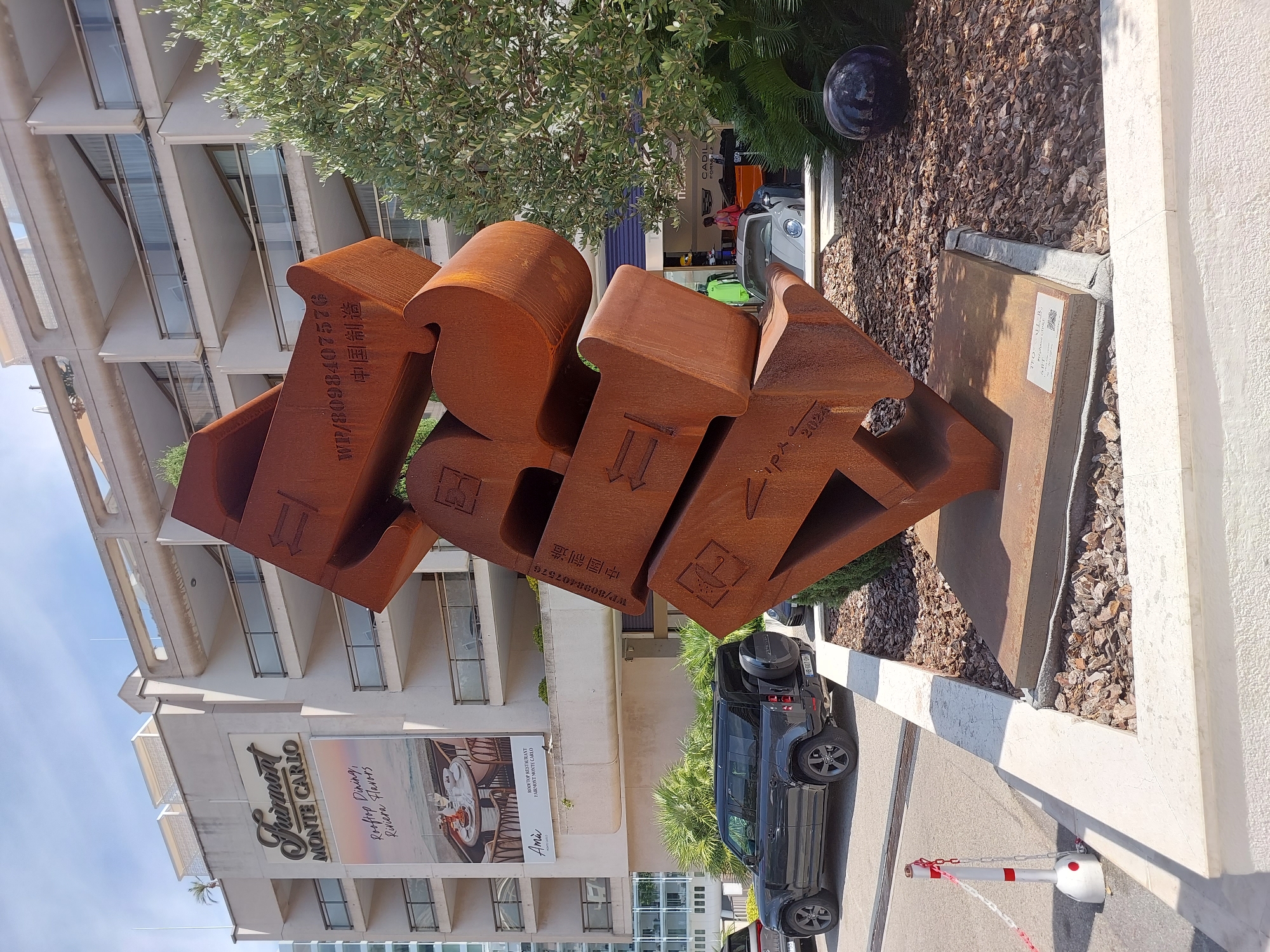 The image shows a large, abstract metal sculpture in front of the Fairmont Monte Carlo hotel in Monaco. The sculpture is made of rusted metal and has an intricate, stacked design. It is located on a street corner with a sidewalk and a landscaping area with mulch and plants. Several cars are visible, including a dark grey Land Rover Defender parked on the street and a silver Porsche in the background. The Fairmont Monte Carlo hotel is a multi-story building with balconies and is identifiable by its prominent signage. The sky is blue with some clouds, and the scene appears to be during the daytime, suggesting a sunny day. A red and white striped stanchion with a chain is visible on the sidewalk near the sculpture.