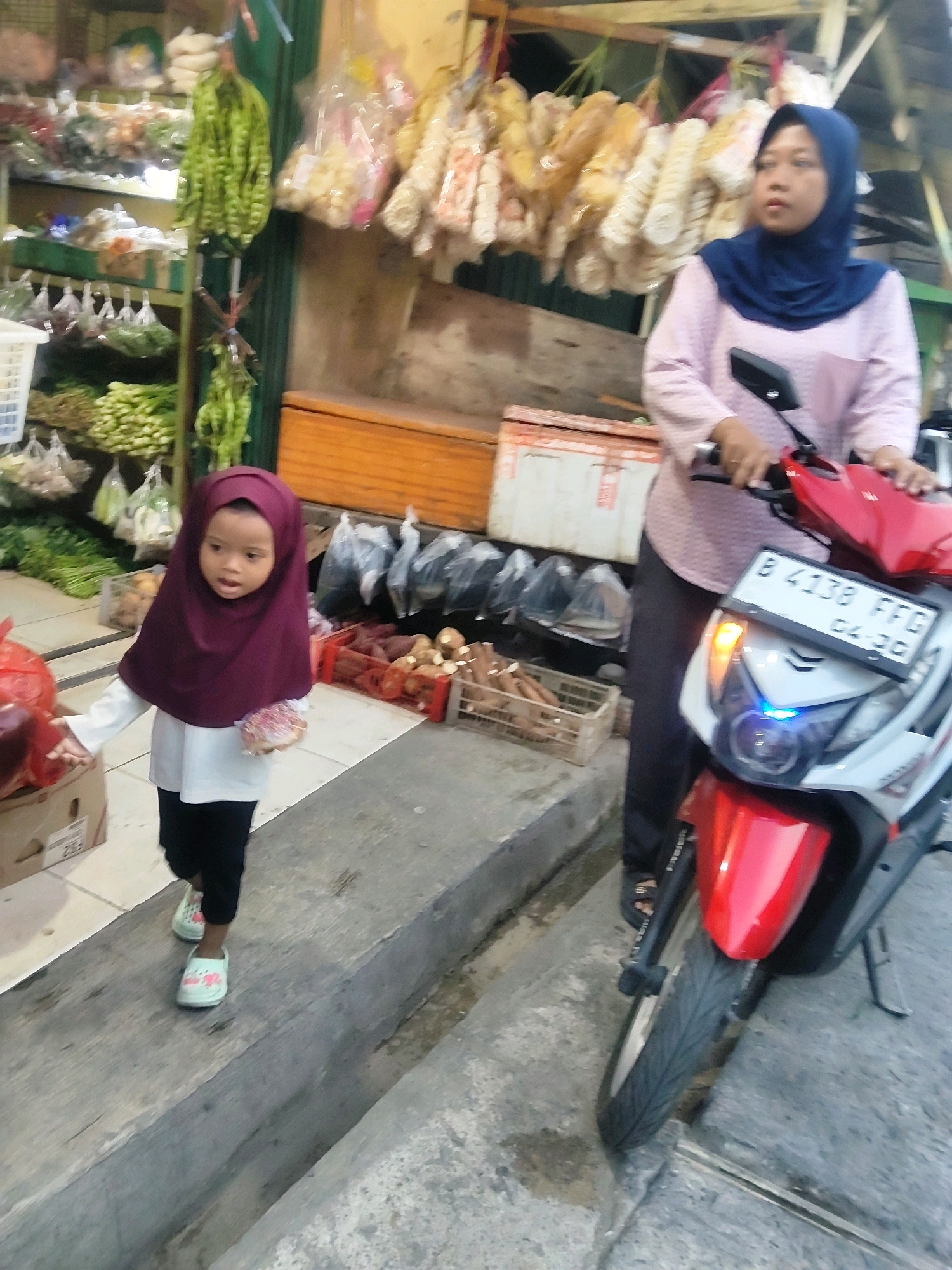 In a street market scene in Mekarmukti, Indonesia, a young child wearing a maroon hijab and white long-sleeved shirt with black pants and light green sandals stands on a concrete path. The child is holding a round, pink object. To the right, a woman wearing a navy blue hijab and a pink patterned long-sleeved shirt is seated on a red and white scooter. She is holding the scooter's handlebars. The scooter has a license plate that reads "B 4138 FFG" and "04-20". Behind the woman, a market stall displays various food items hanging from the ceiling and arranged on shelves. To the left of the child, market stalls with produce, including green vegetables and other packaged goods, are visible. The ground is made of concrete, with a visible drain or gutter.