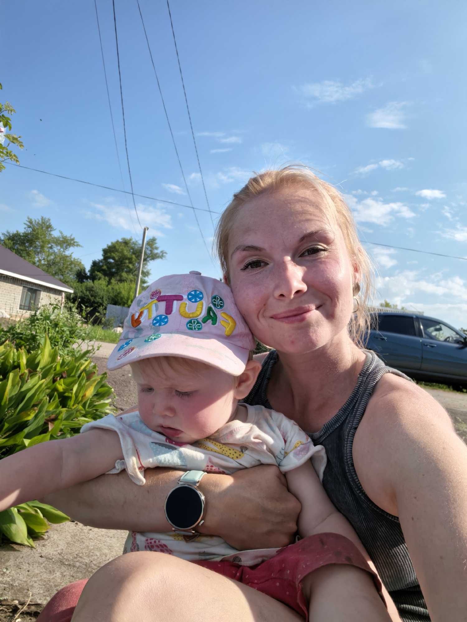 A medium shot captures a woman holding a baby in her arms, both facing the camera in a selfie. The woman, with blonde hair and light skin, smiles warmly. She is wearing a gray, ribbed tank top and gold hoop earrings. The baby, wearing a pink cap with colorful letters and a patterned onesie, looks down and away from the camera. The baby's left arm is visible, with a smartwatch featuring a round, black face and a light blue band on the wrist. Behind them, a suburban setting unfolds under a bright blue sky with scattered clouds. A brick house with a dark roof is visible on the left, surrounded by green foliage. A blue vehicle is parked on the right side of the frame. The overall lighting suggests it is daytime. The image appears to be taken outdoors in a residential area, possibly in Sterlitamak, Russia, given the context.
