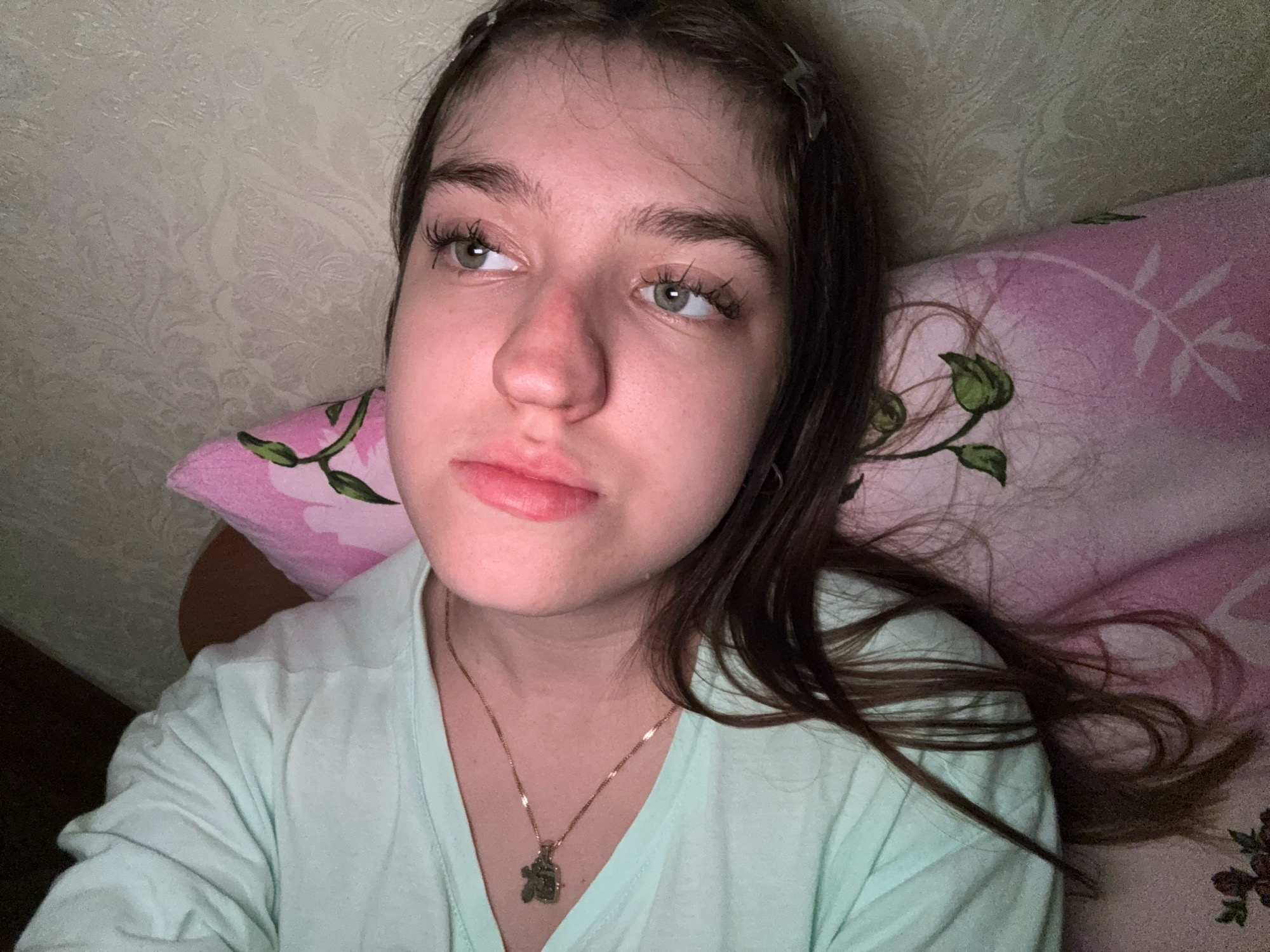 The image is a medium shot of a young person lying down, with their head slightly propped up. They are looking off to the side, with a neutral expression. The person has long, dark brown hair, with some strands falling across their face. They are wearing a light green t-shirt and a gold chain necklace with a small, intricate pendant. The background appears to be a bedroom, with a textured wallpaper on the wall and a pillow with a pink floral pattern. The lighting is dim and appears to be artificial. The image does not provide any specific details that would place it in the city of Zavolzhye, Russia.