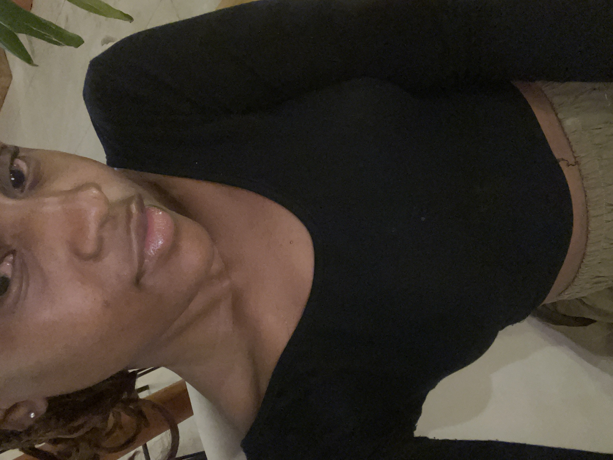 A woman with dark skin and dark curly hair is captured in a close-up, slightly angled shot. She is wearing a black long-sleeved top with a scooped neckline. Her lips are glossy, and she has a small stud earring in her left ear. The background is blurred, but appears to be an indoor setting with hints of furniture and greenery. The image was taken in Gura Top, Nigeria.