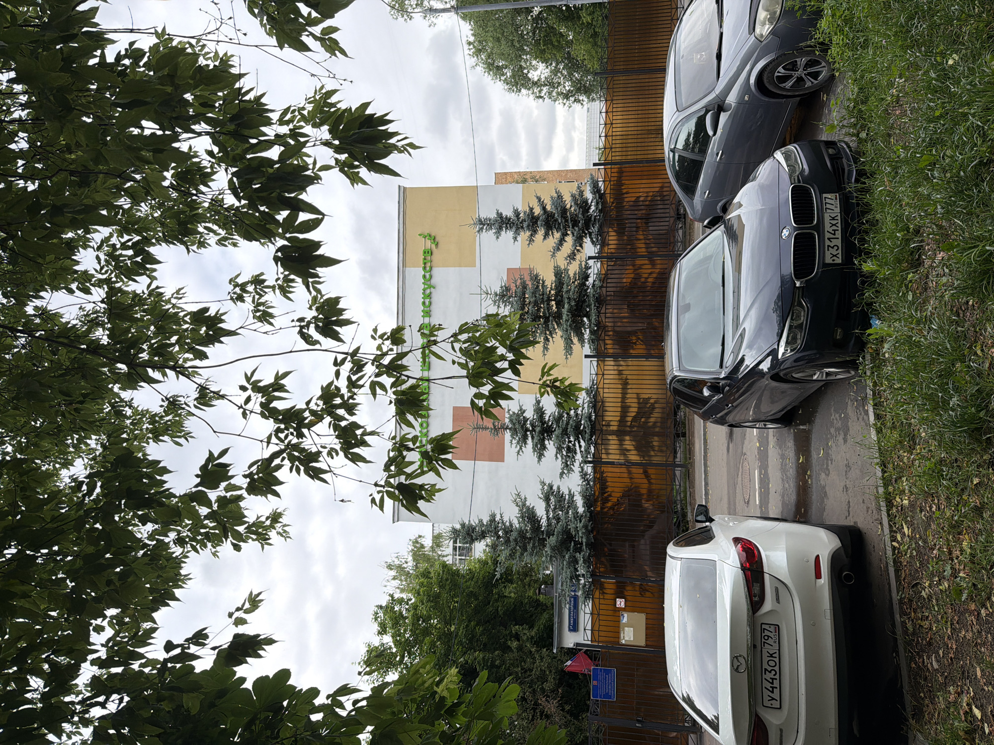 The image shows a street view with trees in the foreground and a building in the background. The building has a sign that reads "ШКОЛА ИСКУССТВ" (Art School). In front of the building, there is a fence and several fir trees. Three cars are parked in front of the fence. The car on the left is a white Mazda 6 with a Russian license plate "У443ОК 797 RUS". The middle car is a dark blue BMW 3 Series with a Russian license plate "Х314ХК 777 RUS". The car on the right is a gray Nissan Almera. The sky is overcast. The ground is wet, suggesting it might have rained recently.