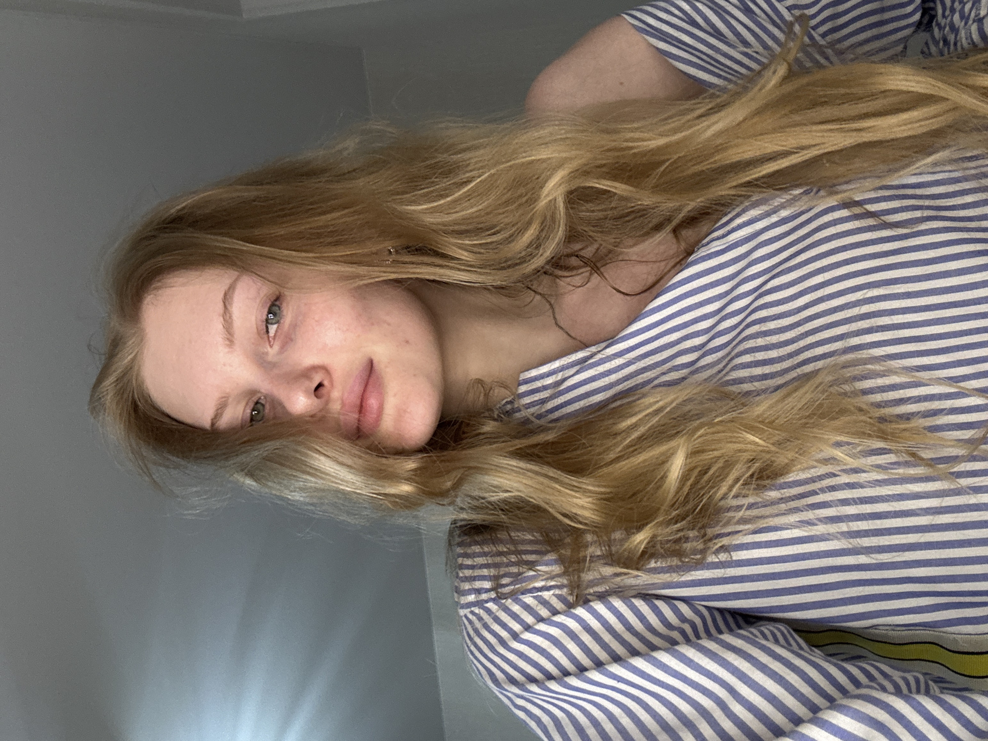 A pale-skinned person with long, light brown hair is looking at the camera. The person is wearing a blue and white striped shirt. The background is a plain, light gray wall. There is no evidence to suggest the location is Perm, Russia.