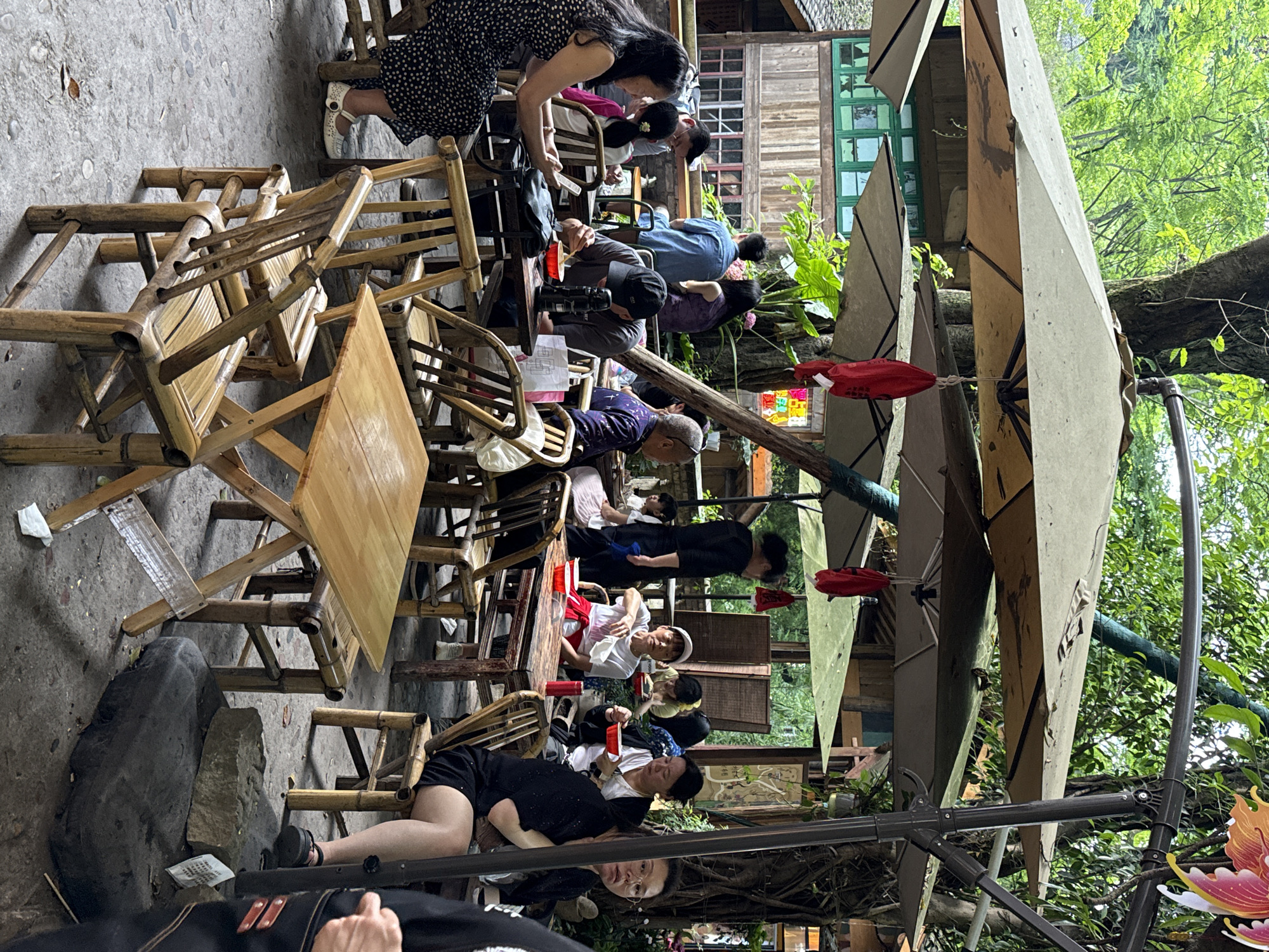 The image shows an outdoor scene in Beibei, China. Numerous people are sitting at bamboo tables and chairs under large umbrellas, likely at a restaurant or cafe. Some individuals are interacting, while others appear to be resting or observing. The environment includes plants, trees, and architectural elements, creating an outdoor setting.