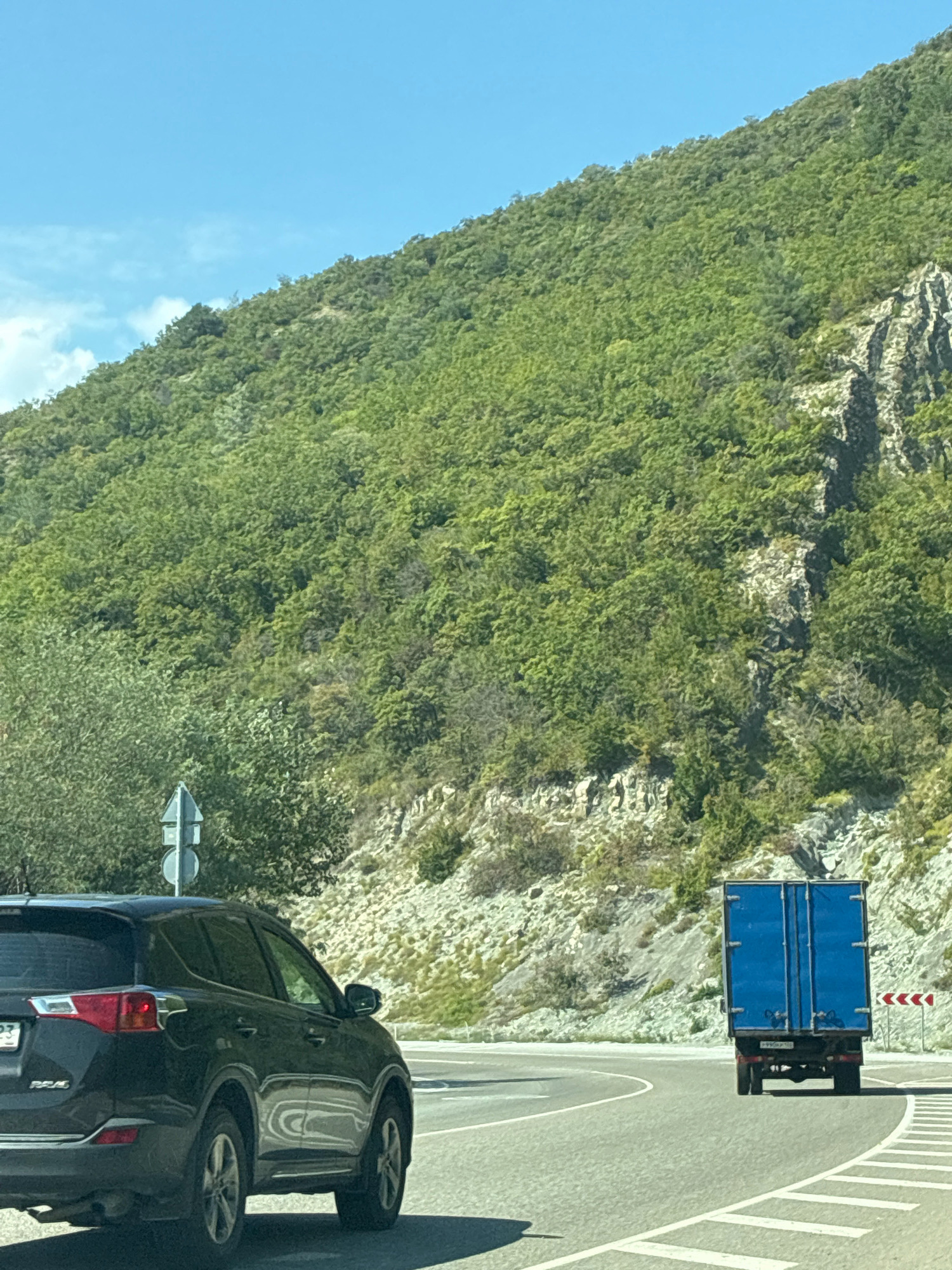 The image captures a road in Shirokaya Schel, Russia, winding around a steep, tree-covered mountainside. The road is paved and marked with white lines. In the foreground, a dark-colored SUV is parked or moving slowly on the left side of the road, facing away from the viewer. Further down the road, a blue truck with a closed trailer is visible, also moving away from the viewer. The mountainside is densely covered in green foliage, with rocky outcrops visible in places. The sky is clear and blue, with a few wispy clouds. A traffic sign with two circular panels is located on the left side of the road, behind the SUV.
