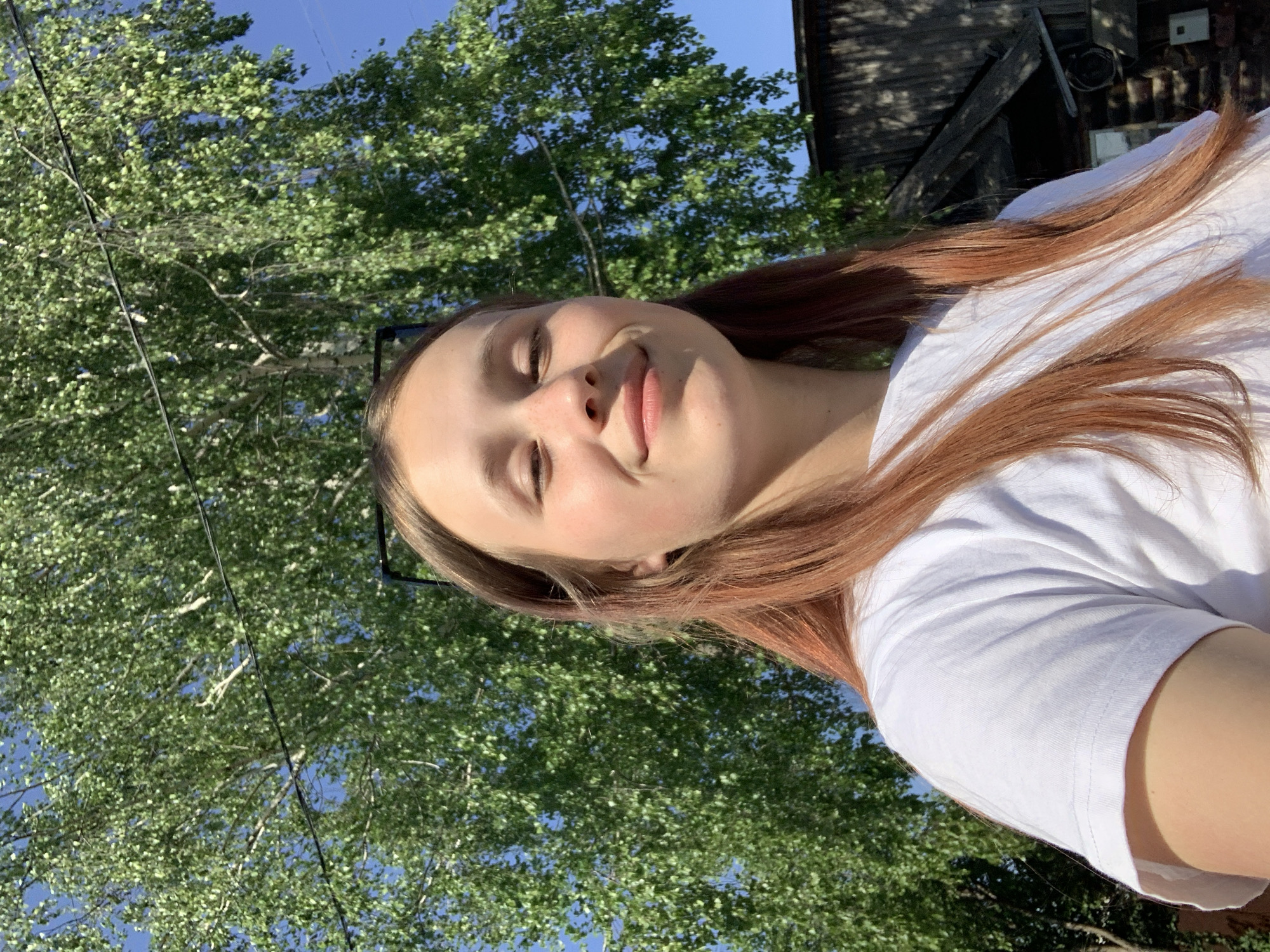 A woman with light skin and red hair is angled in the frame, wearing a white shirt. Behind her is a large tree with green leaves against a blue sky. In the upper right corner of the image is a building. There is no way to determine from the given information that this image was taken in the city of Фоки, Russia.