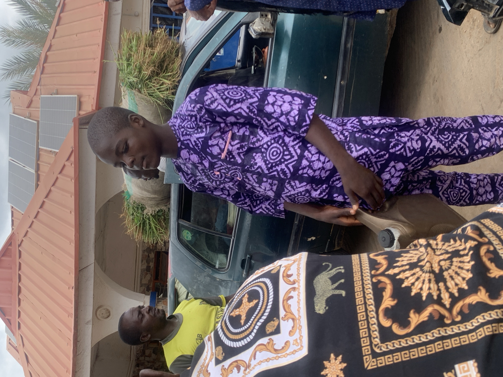 The image shows a group of people gathered near a green car in Akwanga, Nigeria. One person in a purple patterned outfit is holding a tan-colored container. Another individual wears a yellow shirt. A bag filled with straw is on top of the car. A building with solar panels is visible in the background. A man in a shirt featuring a leopard is present as well.