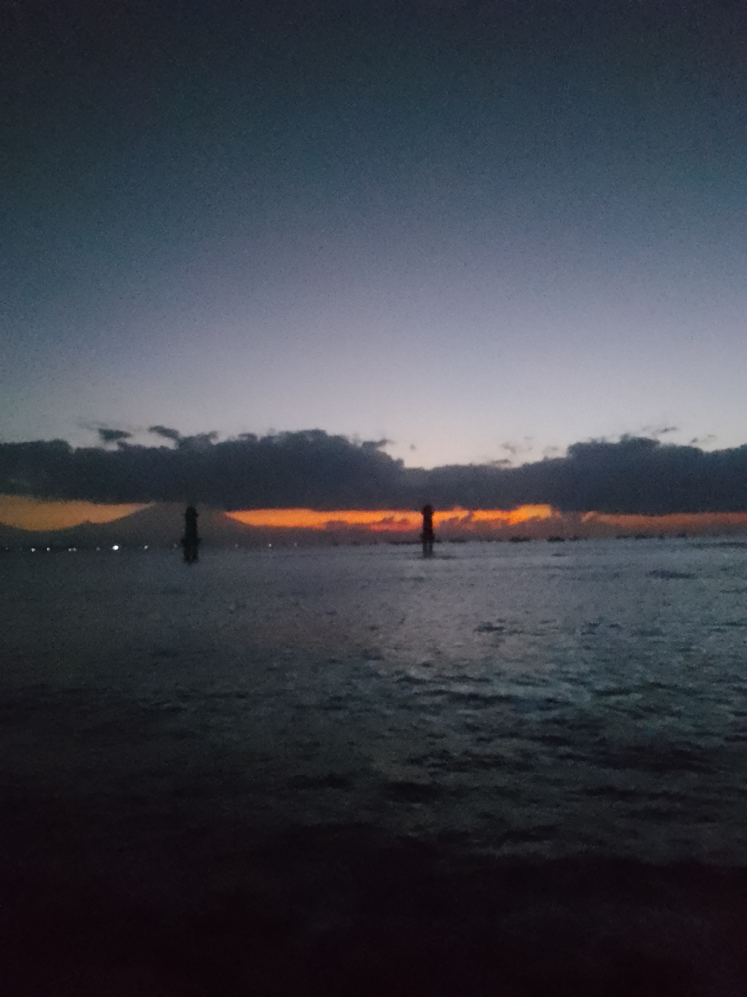 The image depicts a serene seascape at dusk, likely from a coastal area in Denpasar, Indonesia. The sky transitions from a deep navy blue overhead to a soft gradient of pale blue and hints of purple closer to the horizon. A band of dramatic clouds, tinged with vibrant orange and yellow hues from the setting sun, stretches across the middle of the frame, creating a striking contrast with the darkening sky.
Below the clouds, the ocean reflects the twilight ambiance. Gentle waves create subtle ripples on the surface, with a darker, almost silhouetted foreground suggesting the viewer is positioned on a shoreline or a structure close to the water. In the distance, silhouetted against the bright horizon, are what appear to be landforms, possibly mountains or islands, with a few scattered lights indicating settlements. A prominent, dark, solitary figure, appearing to be a structure like a lighthouse or a tower, stands in the water, adding a focal point to the scene. The overall atmosphere is one of tranquility and natural beauty as daylight fades.