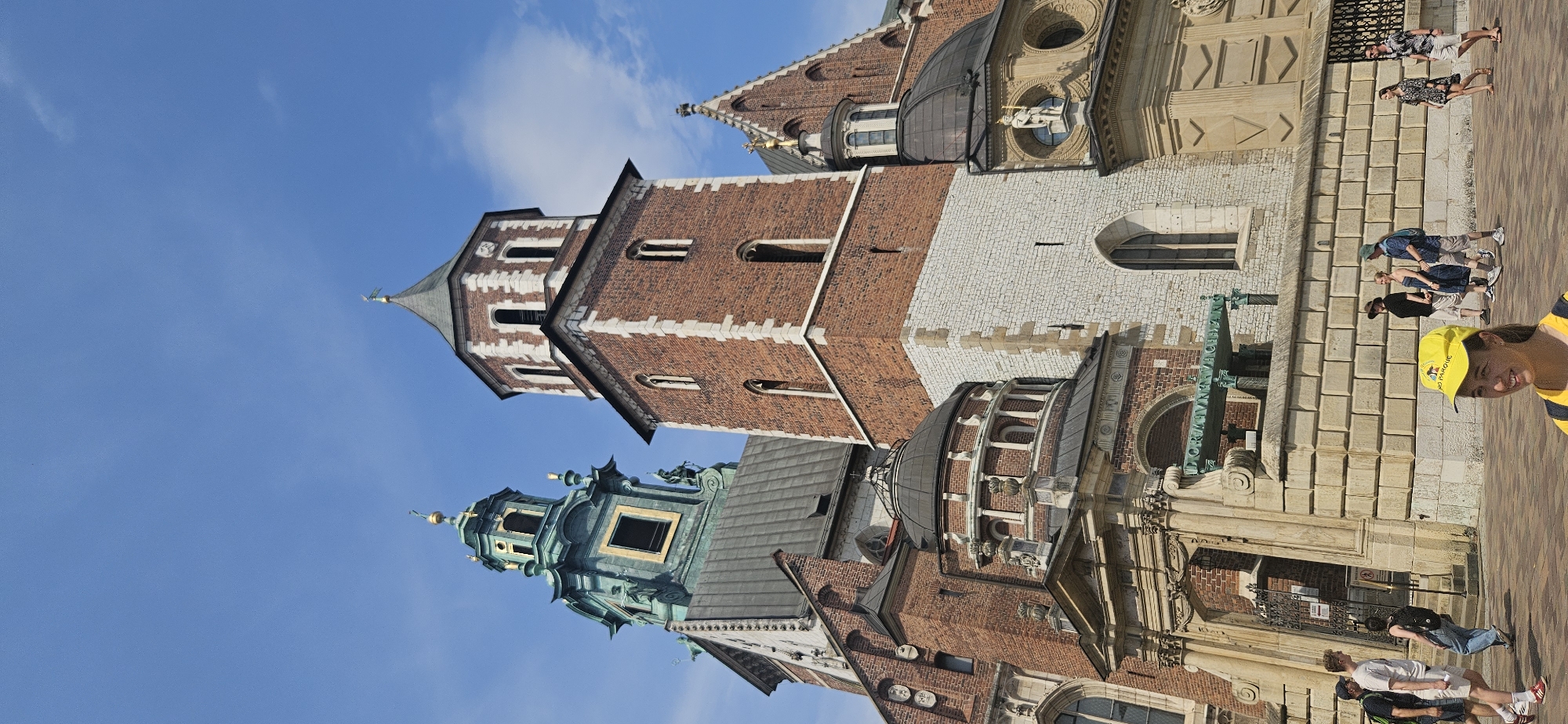 The image shows a daytime exterior view of Wawel Cathedral in Krakow, Poland. The cathedral features several distinct architectural elements, including a tall, red brick tower with white stone accents, a green-domed tower with gold embellishments, and multiple dark domes. In the foreground, a group of people are walking on a paved area in front of the cathedral. A young woman wearing a yellow baseball cap is prominently featured in the lower right portion of the image, looking towards the left. Other individuals are dressed in casual summer attire. The sky is blue with scattered white clouds. The overall impression is of a tourist destination on a clear day.