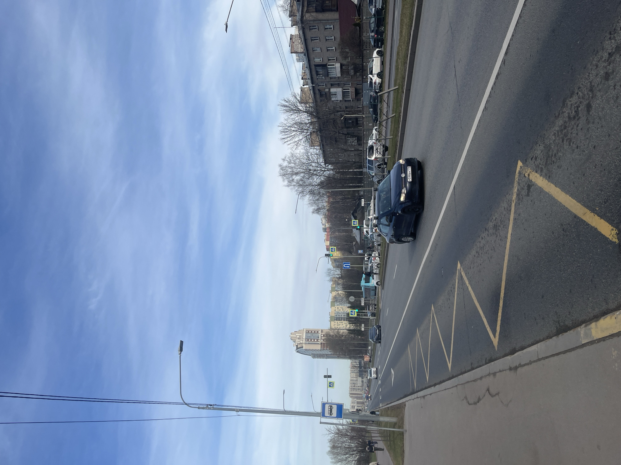 The image depicts a street scene in Saint Petersburg, Russia. A four-lane road stretches into the distance, with vehicles in both directions. A dark-colored Volkswagen Polo, license plate "Р598ВН 178", is in the nearest lane, moving away from the viewer. Yellow zigzag lines mark the edge of the lane, indicating a bus stop. The sidewalk is to the right of the road. On the left of the road, there are apartment buildings and trees. Streetlights line the road. The sky is blue with streaky clouds. A bus stop sign is visible in the distance.