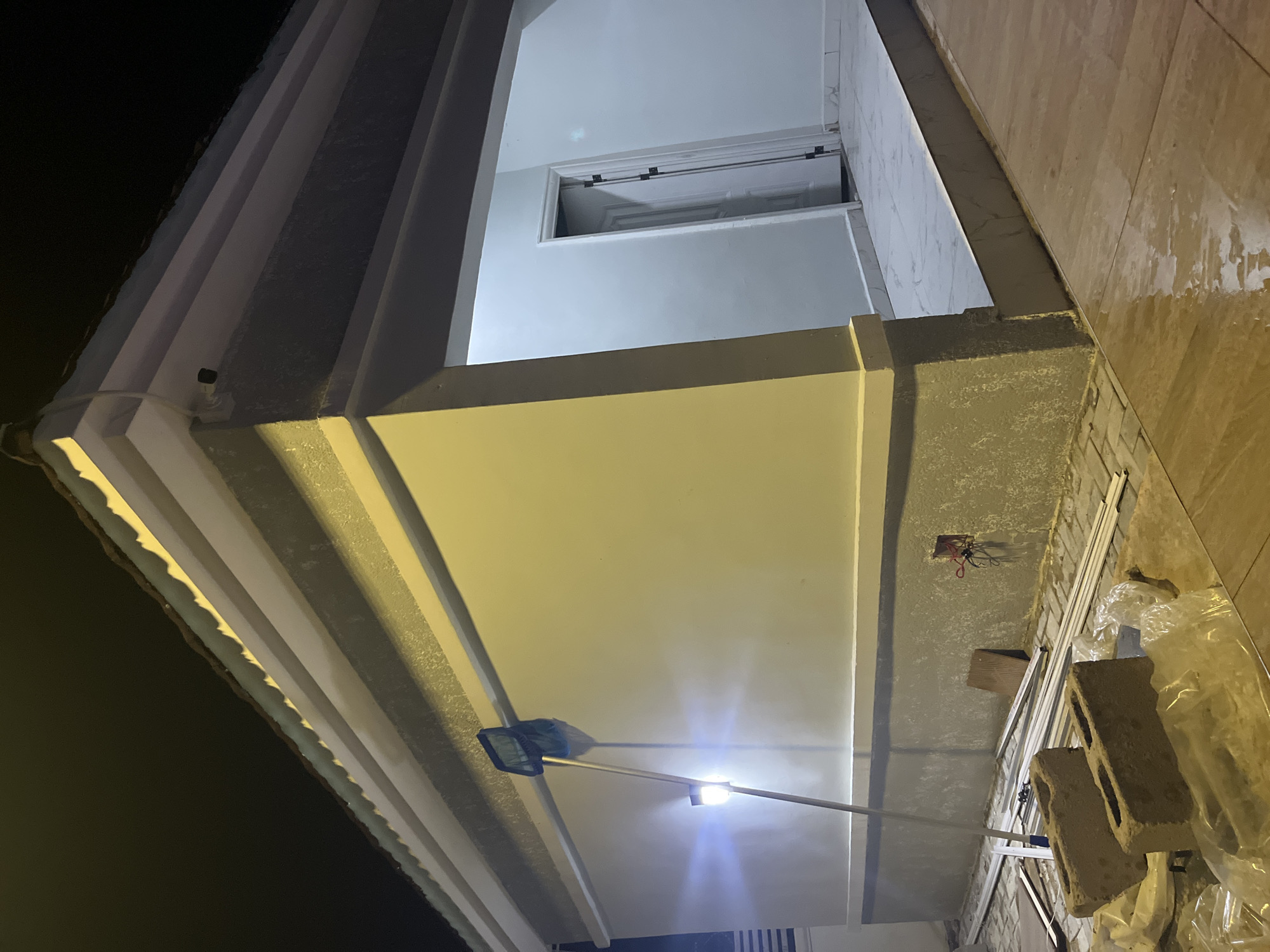 The image shows the exterior of a building at night, likely a residential property under construction or renovation in Kason, Nigeria. The building has a modern design with a light-colored facade and a dark-colored roof. A white-colored door is visible, leading into the building. There are construction materials scattered around the exterior, including bricks, plastic sheeting, and metal pipes. A broom with a blue net is leaning against the wall. A light fixture is illuminating the wall. The ground appears to be paved with tiles and is wet, suggesting recent rain or cleaning. The overall impression is one of a work in progress, with the building being prepared for occupancy or further development. The scene is dimly lit, with artificial lights providing the primary illumination.