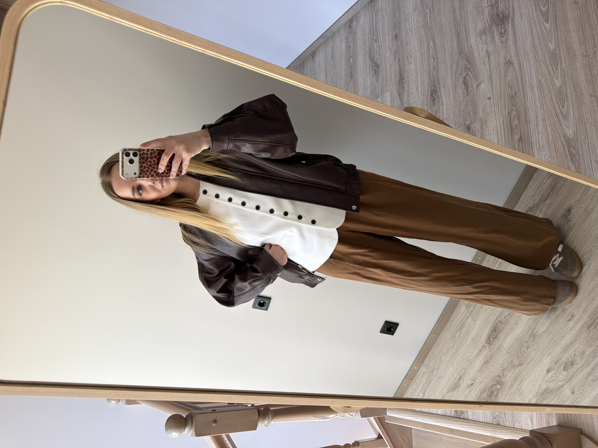 A young woman is captured in a mirror selfie, showcasing her outfit indoors in what appears to be a residential setting in Dreiliņi, Latvia. She has long, straight blonde hair and holds a smartphone with a leopard print case, which partially obscures her face as she takes the photo. Her visible eye has a neutral expression.
She is dressed in a fashionable ensemble consisting of a dark brown, possibly faux leather, bomber jacket with an elasticized hem and cuffs. Beneath the jacket, she wears a crisp white collared top with a line of small black buttons down the center. Her lower half is clad in wide-leg, camel-brown trousers that fall loosely over her footwear. She completes her look with light brown or grey sneakers featuring white laces.
The photo is taken in a large rectangular mirror with a light wooden frame. The room features light-colored walls and a light-toned wooden floor with a visible grain pattern. Two black electrical outlets are discreetly placed on the wall behind her. Part of a light wooden chair or stool with turned legs is visible in the reflection at the bottom of the mirror. The lighting suggests it is daytime, providing a clear view of the interior space. No discernible text is visible in the image.