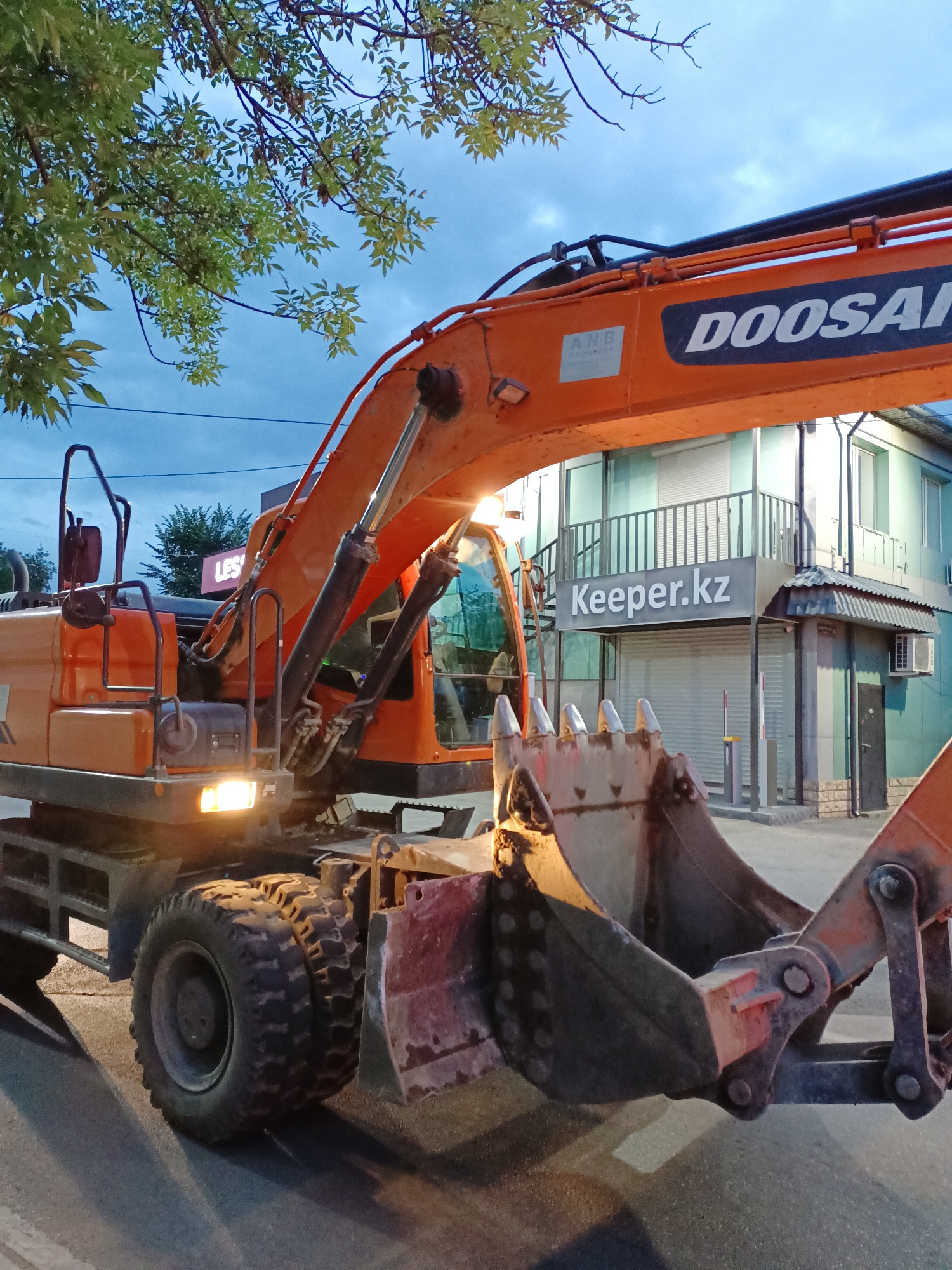 An orange Doosan excavator is parked on an asphalt road at dusk in Almaty, Kazakhstan. The excavator's arm is extended, and its bucket is lowered to the ground. A building with a sign that reads "Keeper.kz" is visible in the background, along with a tree with green leaves. The sky is cloudy and dark blue. The excavator's headlights are on, illuminating the road in front of it.