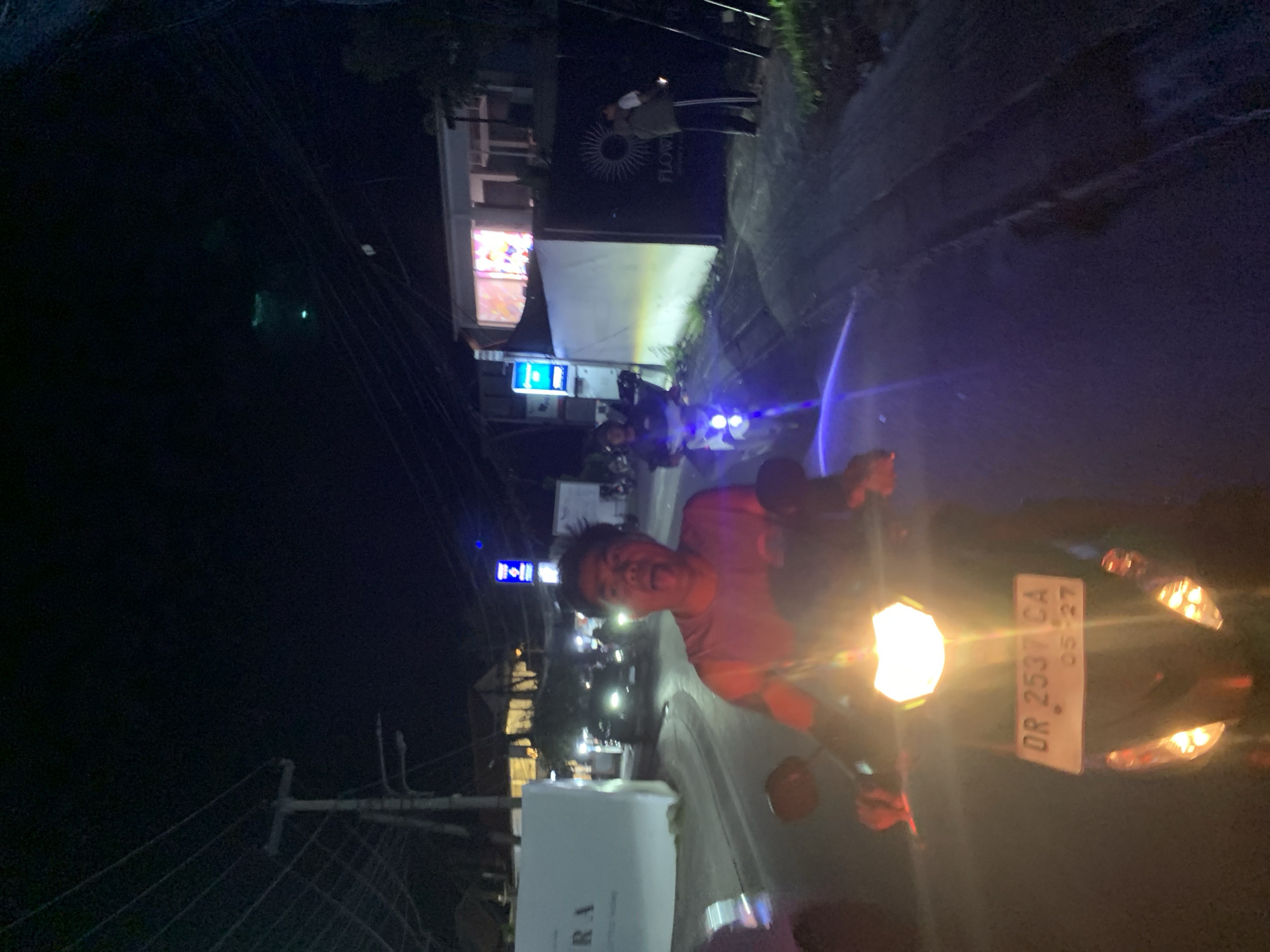 The image shows a man riding a motorbike at night in North Kuta, Indonesia. The man is fair-skinned and wearing a pink shirt. He is smiling and looking at the camera. The motorbike has a license plate that reads "DR 2537 CA" and a sticker indicating the month of May and the year 2027. The motorbike's headlight is bright, illuminating the road ahead. The environment includes buildings, streetlights, and power lines. There are other motorbikes and people visible in the background.