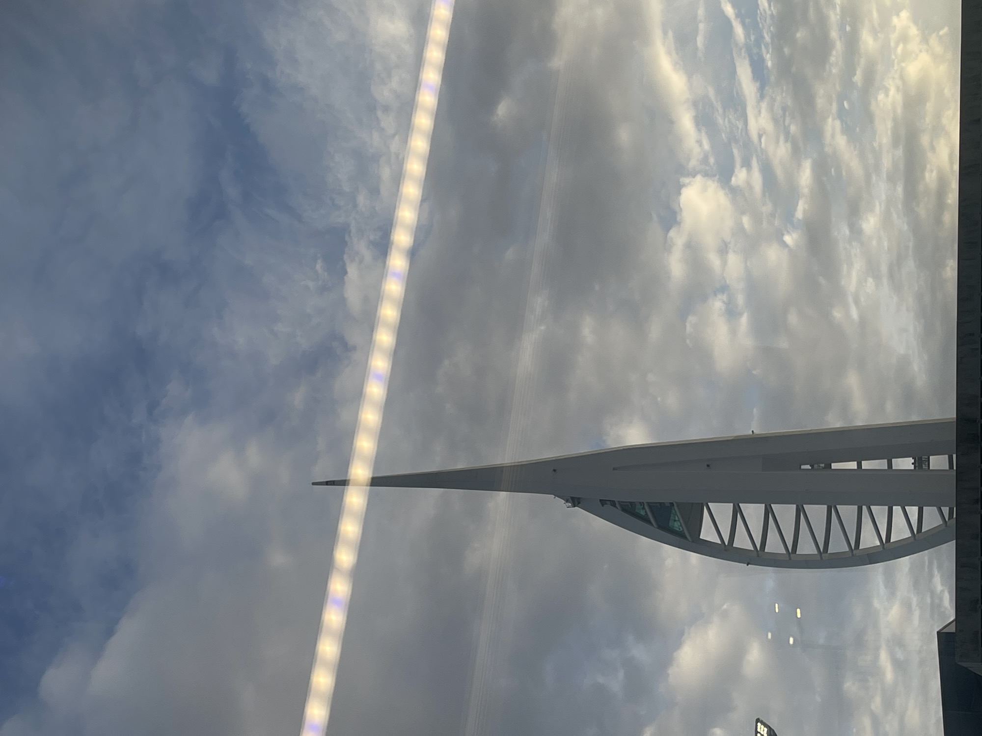 The image displays a prominent architectural landmark, the Spinnaker Tower in Portsmouth, United Kingdom, set against a cloudy sky. The tower is a tall, slender white structure with a distinctive pointed top and a broad, curved base featuring a geometric lattice design.
The sky is a dominant feature, filled with a mix of grey and white clouds, some appearing bright from sunlight while others are darker and more diffused. The overall impression is of a partly cloudy or overcast daytime scene, possibly late afternoon, given the quality of light.
The scene appears to be viewed through a reflective surface, likely a window. This is indicated by a bright, linear light fixture reflecting prominently across the left side of the frame. There are also other fainter, indistinct reflections visible, especially towards the bottom right. No people or specific activities are depicted. While some letters, possibly "TSW," are faintly visible on a horizontal bar of the tower's structure, they are not clearly legible due to the distance and angle. The image captures the modern architecture of the tower juxtaposed against the natural elements of the sky.