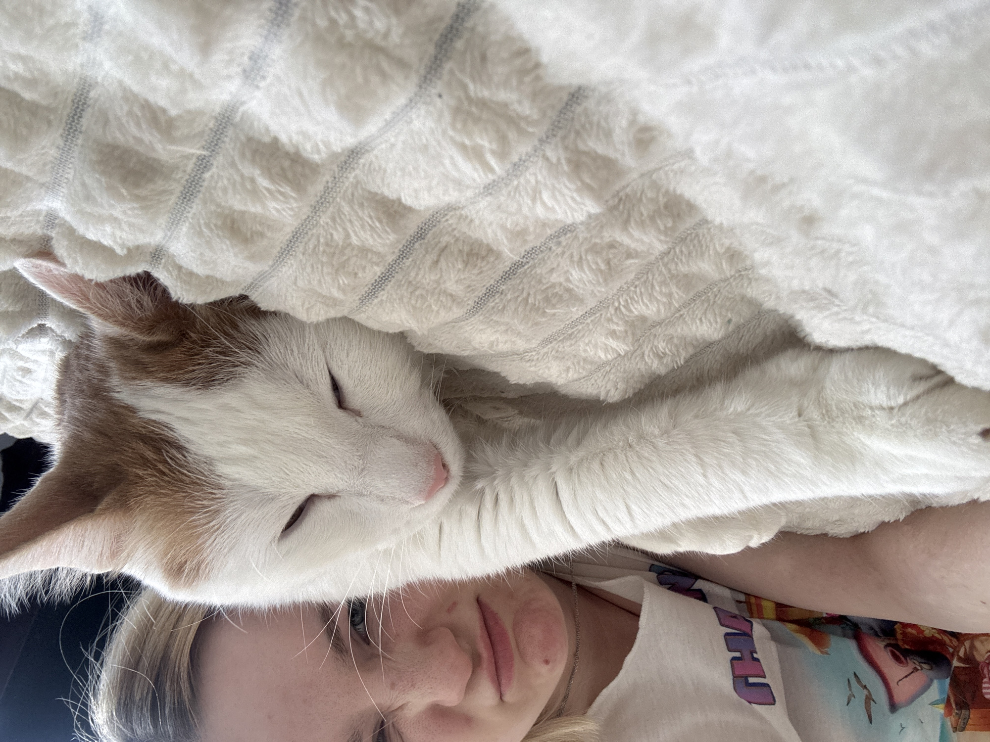 A close-up shot depicts a fair-skinned person with blonde hair and a white and orange cat nestled beneath a white textured blanket. The person's face is visible, with closed eyes, freckles, and a neutral expression. The cat is also lying down with its eyes closed and paws extended. The background is indistinct. The location is unknown, and Vsevolozhsk, Russia, cannot be confirmed from the image.