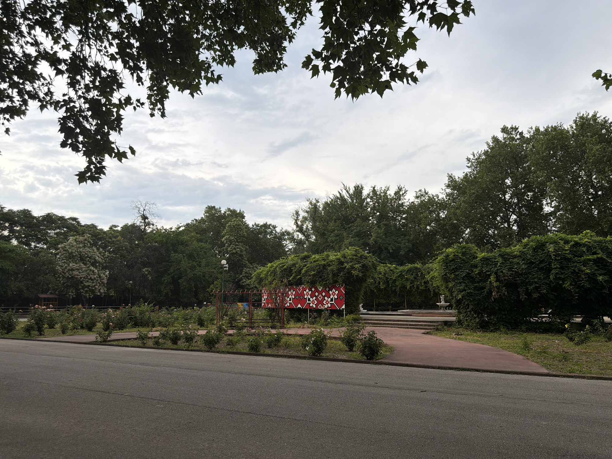 The image depicts a park area in Pazardzhik, Bulgaria, likely on a cloudy day. In the foreground, there is a paved road and a walkway bordering a garden area with numerous rose bushes. Behind the garden, a large, ornate red and white patterned structure is visible, partially covered in greenery. Further into the park, there are more trees, a fountain, and what appears to be a tiered seating area. The sky is overcast with scattered clouds. The scene is devoid of people.
