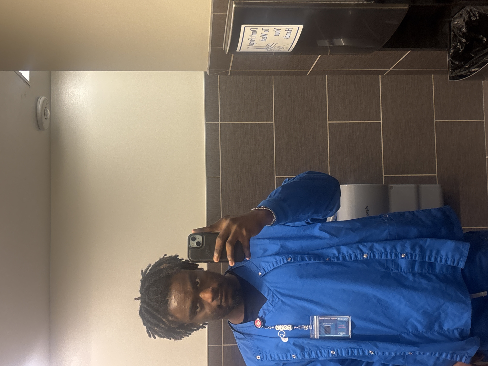 A person is taking a selfie in a bathroom. The person has dark hair and is wearing a blue button-down shirt, and a lanyard with an ID badge. The bathroom has light beige walls on the left side, and gray rectangular tiles with light grout on the right side. A paper towel dispenser is mounted on the wall, as well as a trash can. A toilet paper dispenser is visible in the background. Without further information, pinpointing the location of this bathroom in Grand Prairie is not possible.
