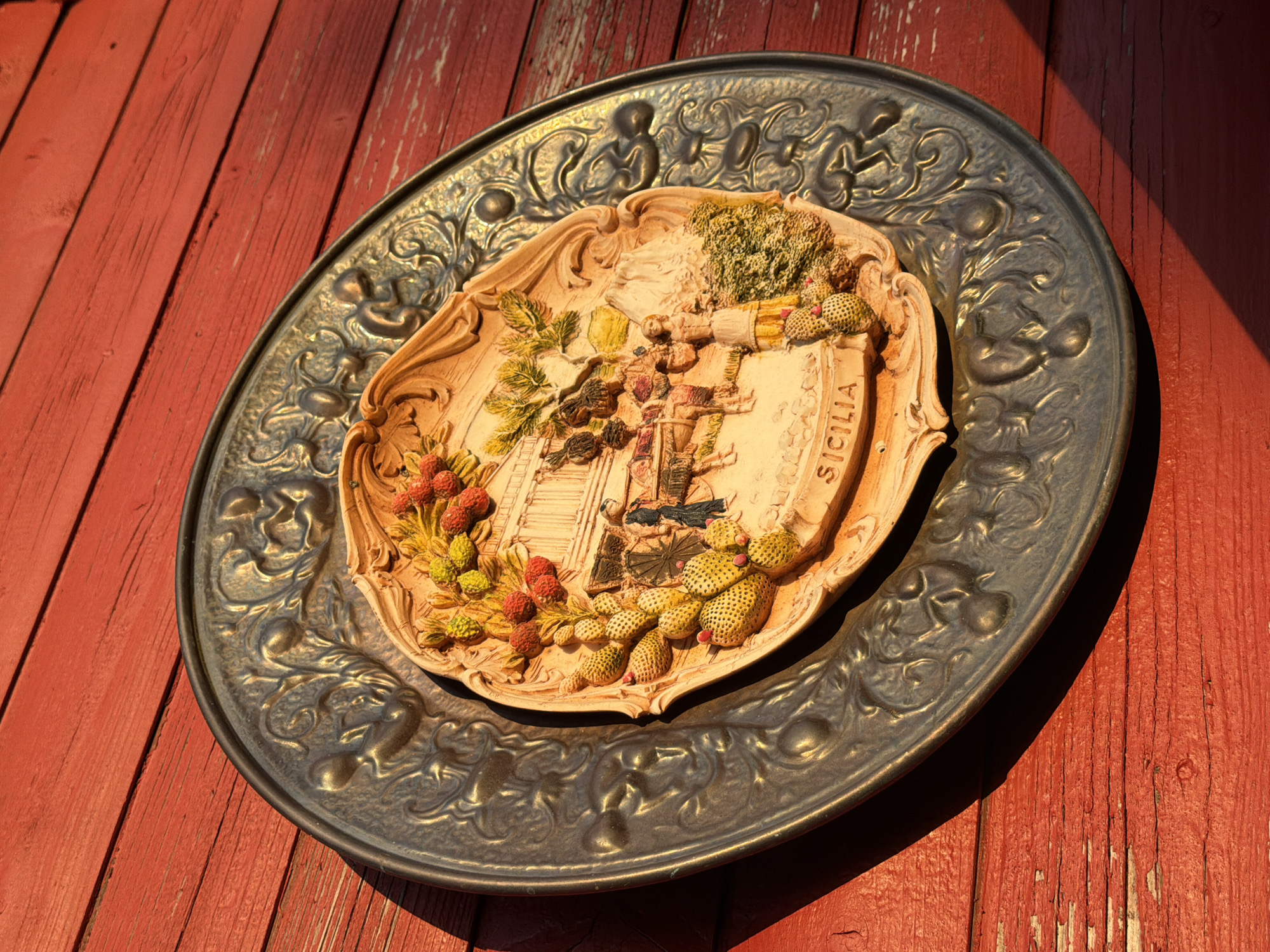 A circular, decorative plate with a raised relief scene is mounted on a weathered red wooden wall. The plate has a dark, ornate outer rim with embossed floral and scrolling patterns. The inner section of the plate is lighter in color and depicts a scene with multiple figures in classical attire, some on horseback. There are also depictions of cactus plants with red and yellow fruit, greenery, and a mountainous landscape in the background. The word "SICILIA" is embossed in the lower portion of the inner relief. The red wooden wall has visible horizontal planks with peeling paint and is illuminated by directional sunlight casting shadows. The location in the city of Bayreuth, Germany, is not discernible from the image.