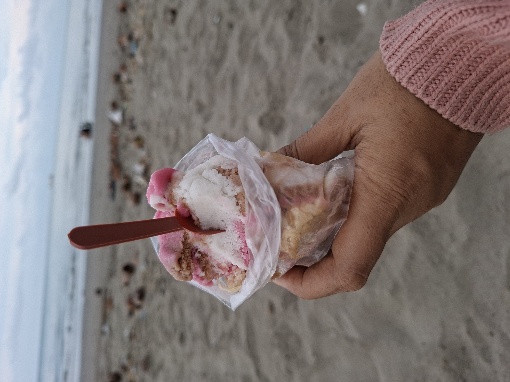A person in a pink knit sweater holds a plastic bag of shaved ice with a brown plastic spoon inserted into it. The shaved ice appears to be layered, with white, pink, and light brown hues visible. The scene is captured outdoors, with a sandy beach as the background. The ocean and cloudy sky are partially visible in the distance. The location is possibly in the coastal city of Lhokseumawe, Indonesia.